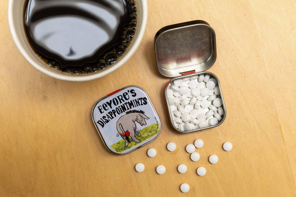 Small tin with white pills, a cup of coffee, and a decorative label on a wooden surface.