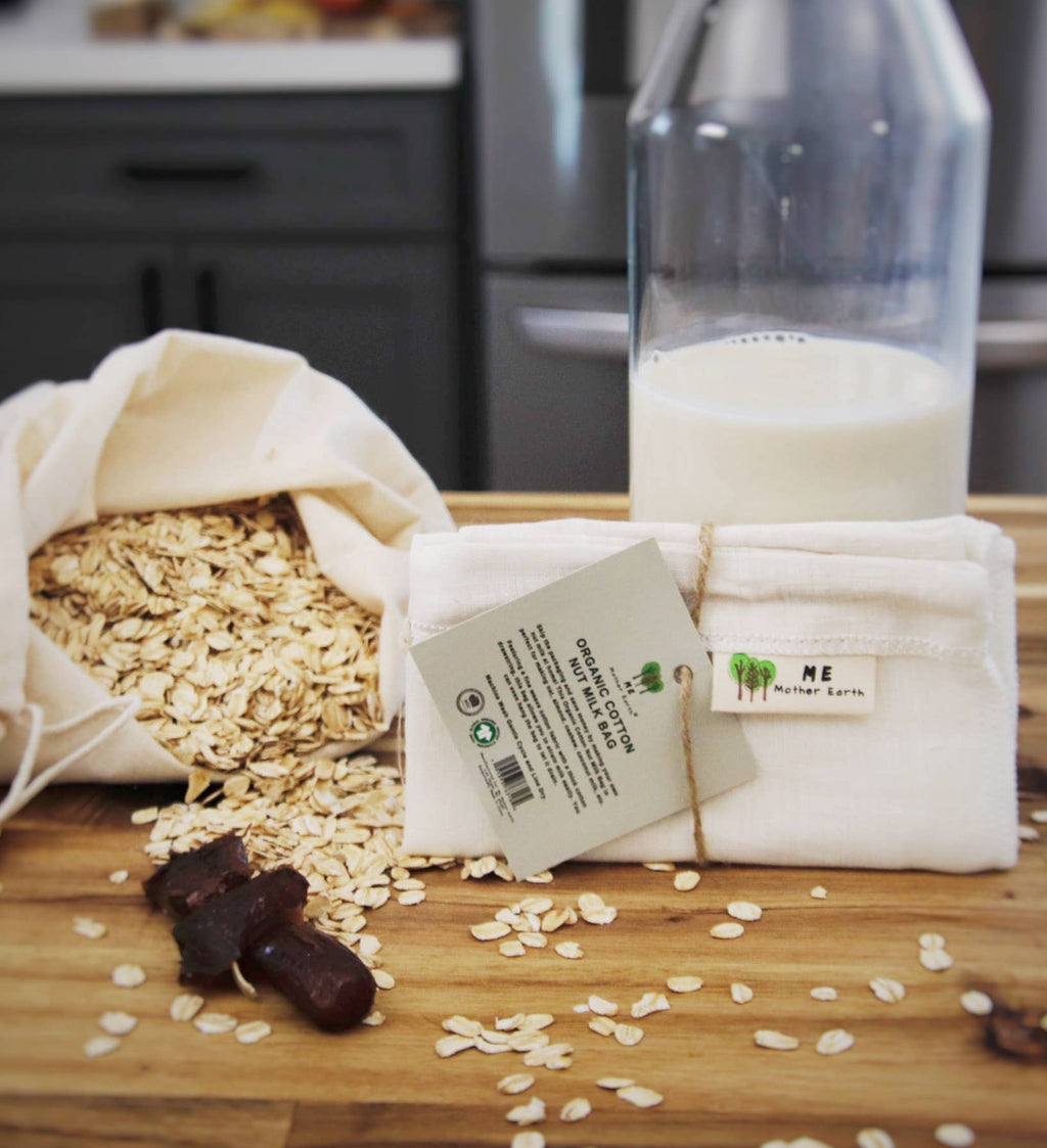 A folded white cloth bag of oats with a paper tag in front, alongside spilled oats, a glass bottle of milk, and a few dates on a wooden tabletop in a kitchen setting.