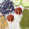 Two round lollipops on sticks with dried fruits, zucchini slices, and herbs on a white surface.