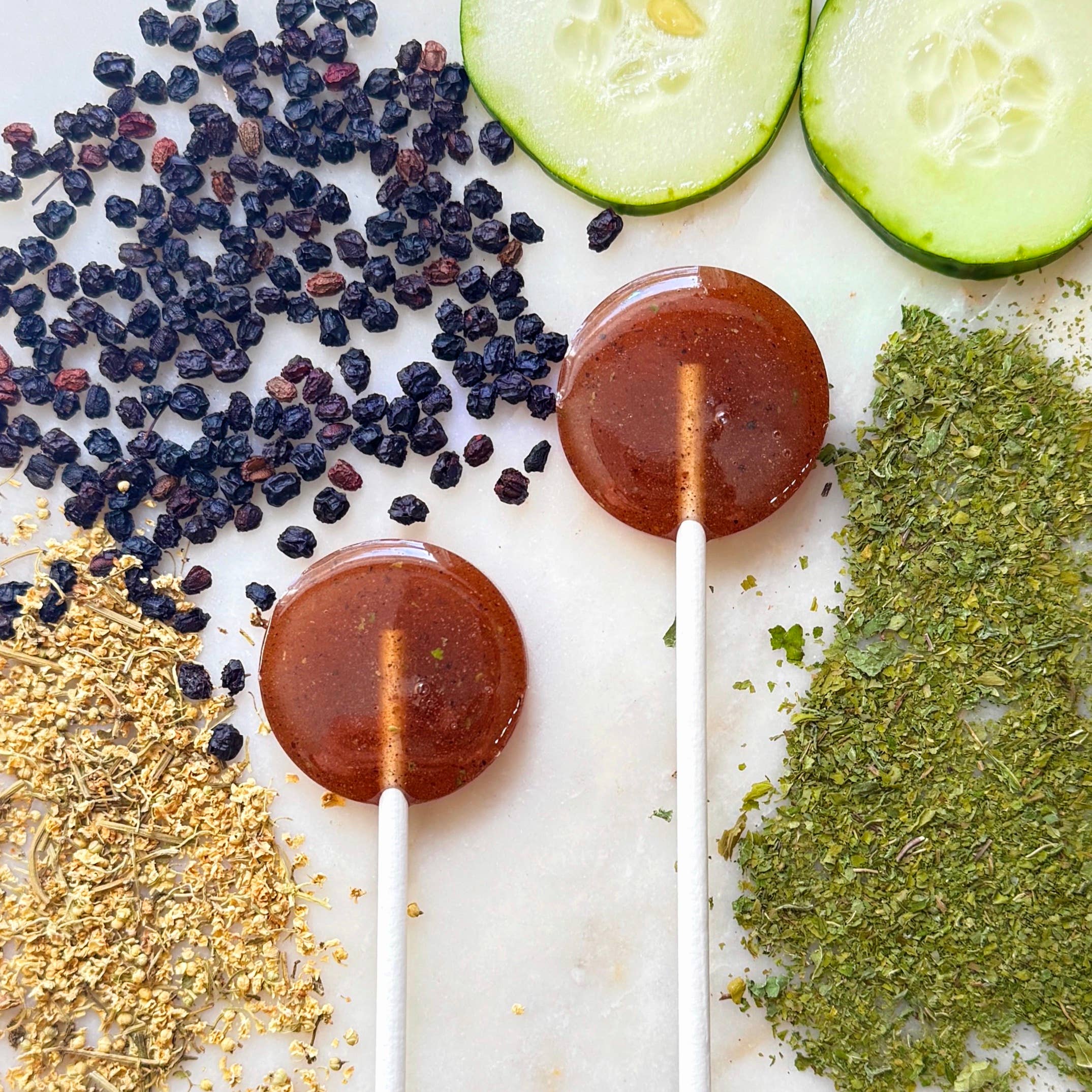 Two round lollipops on sticks with dried fruits, zucchini slices, and herbs on a white surface.