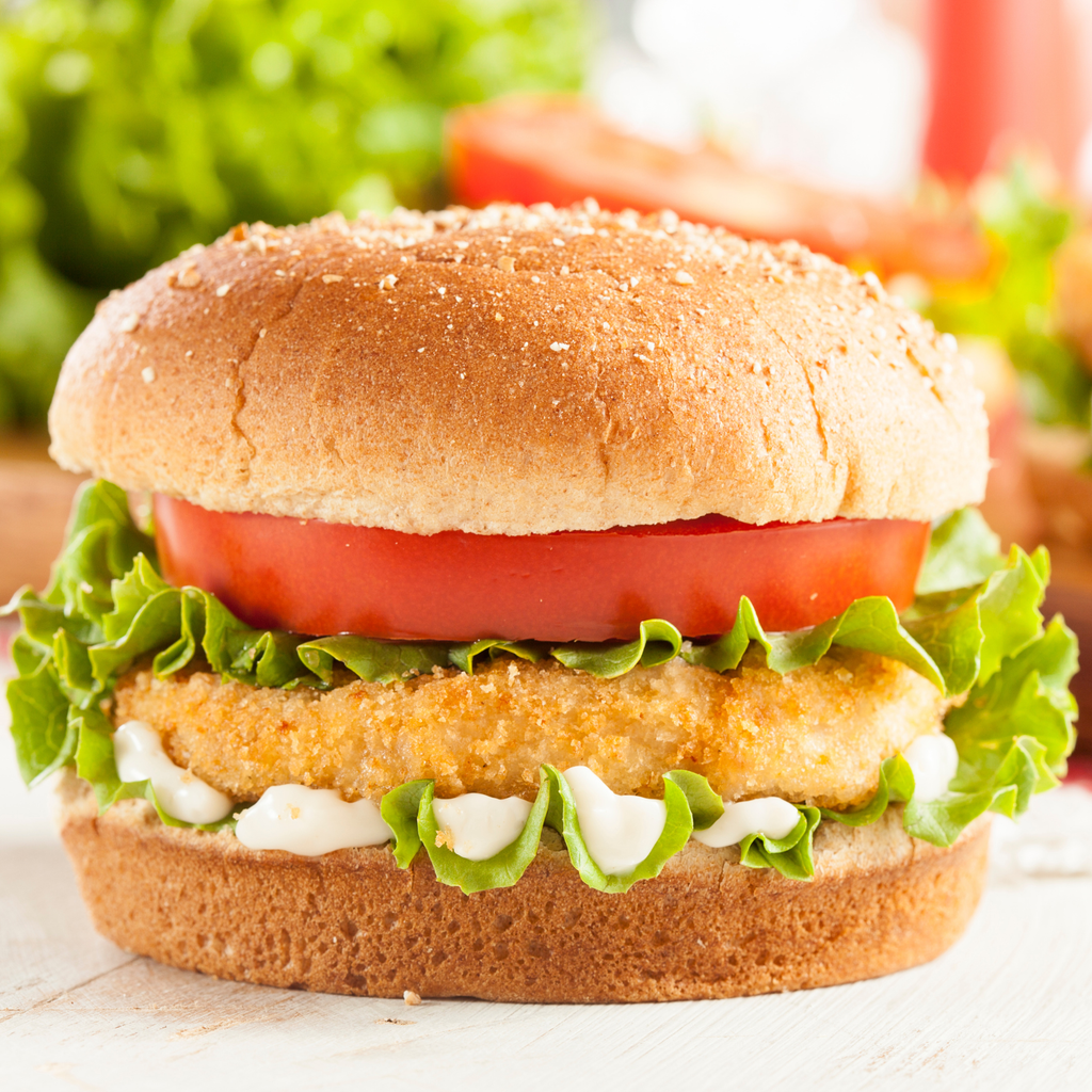 A close-up of a gourmet vegan chicken patty with a golden-brown bread bun, a thick slice of red tomato, fresh green lettuce, a crispy fried patty, and white sauce on a white wooden surface.