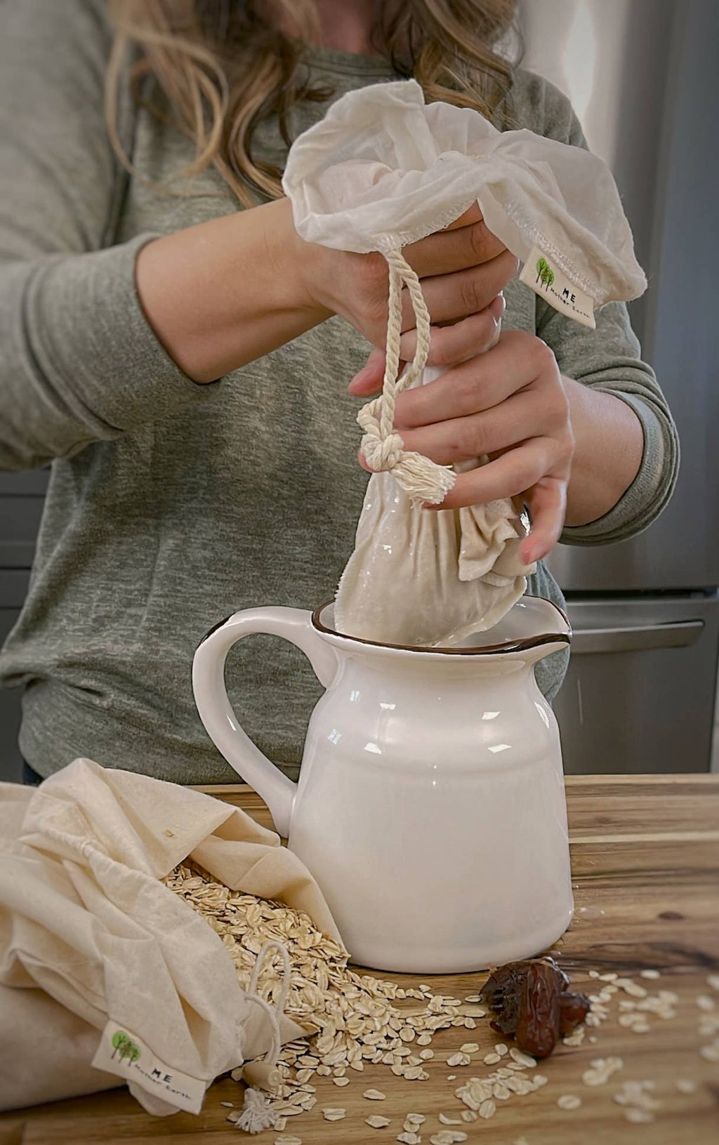 A person wearing a gray long-sleeve shirt demonstrates how to use a reusable cotton drawstring coffee bag to filter coffee from a white ceramic pitcher, with loose oats and a coffee bean on a wooden table.