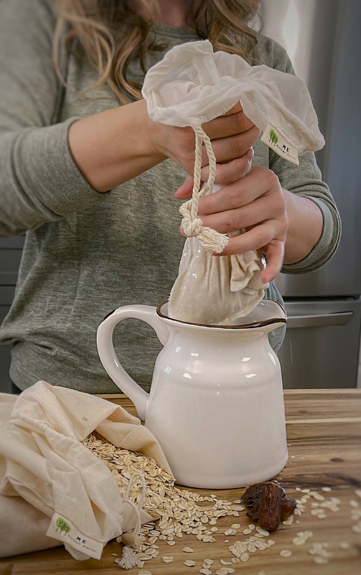 A person wearing a gray long-sleeve shirt demonstrates how to use a reusable cotton drawstring coffee bag to filter coffee from a white ceramic pitcher, with loose oats and a coffee bean on a wooden table.
