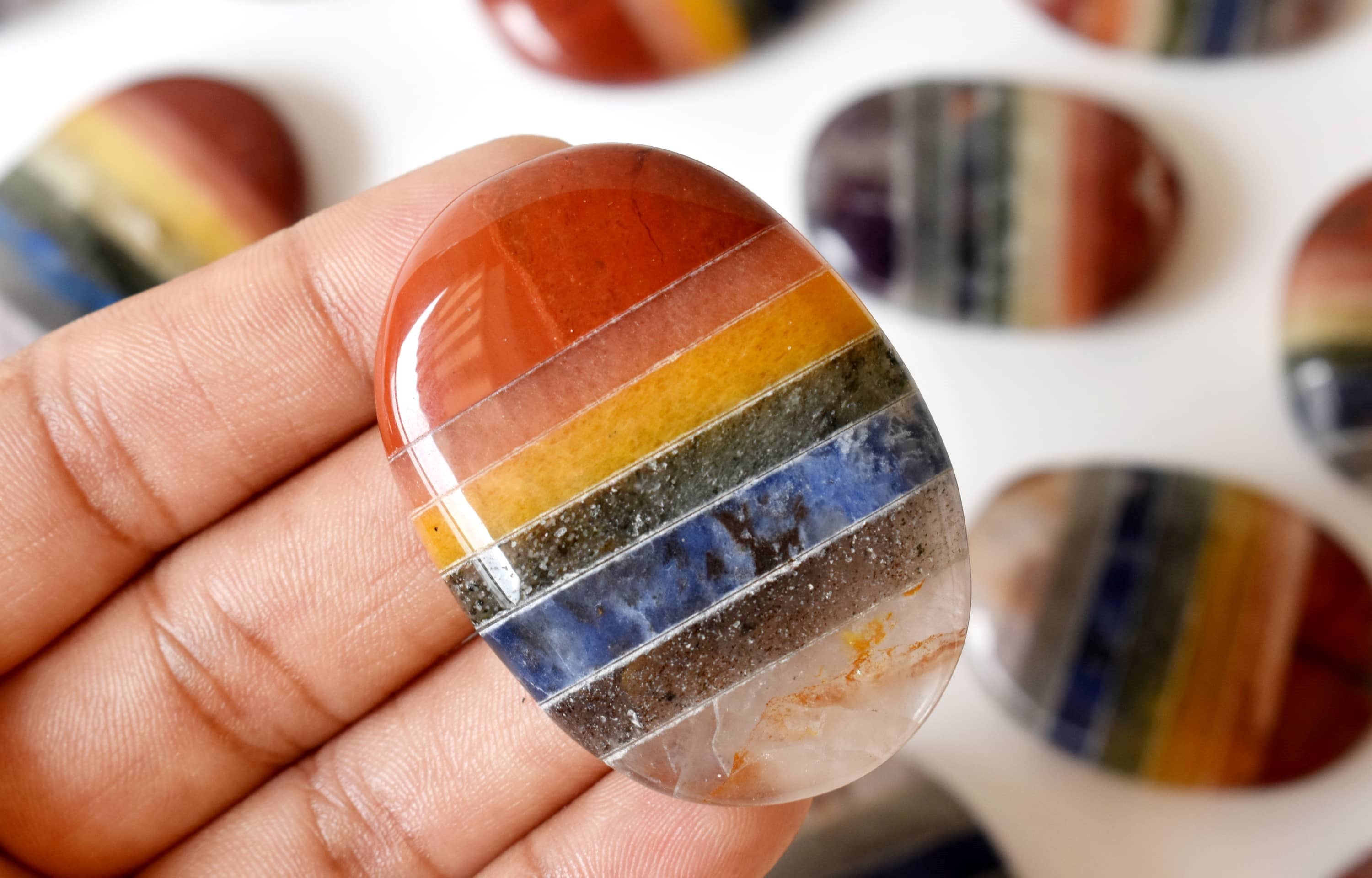 Close-up of a hand holding a rainbow-colored stone with blurred stones in the background.