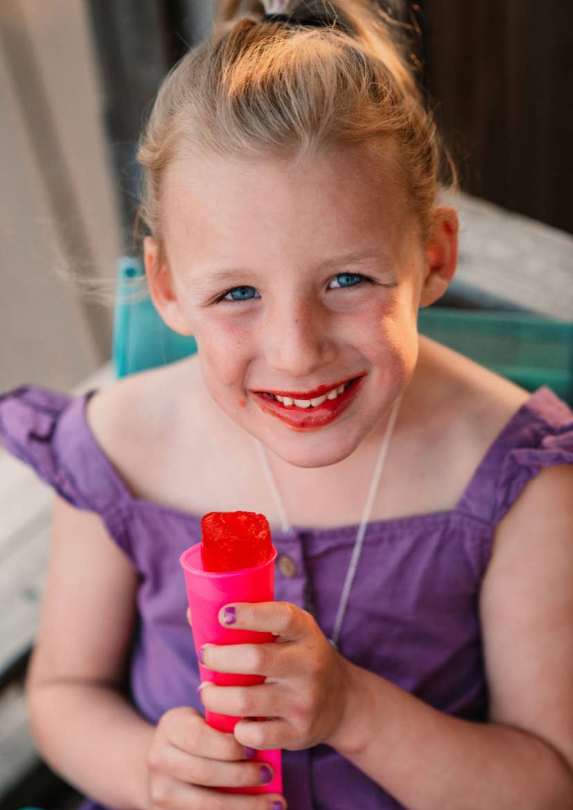 Young girl in a purple dress holding a pink push pop, smiling.
