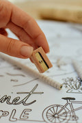 Hand holding a small wooden pencil sharpener with a blurred background of a coloring book page.