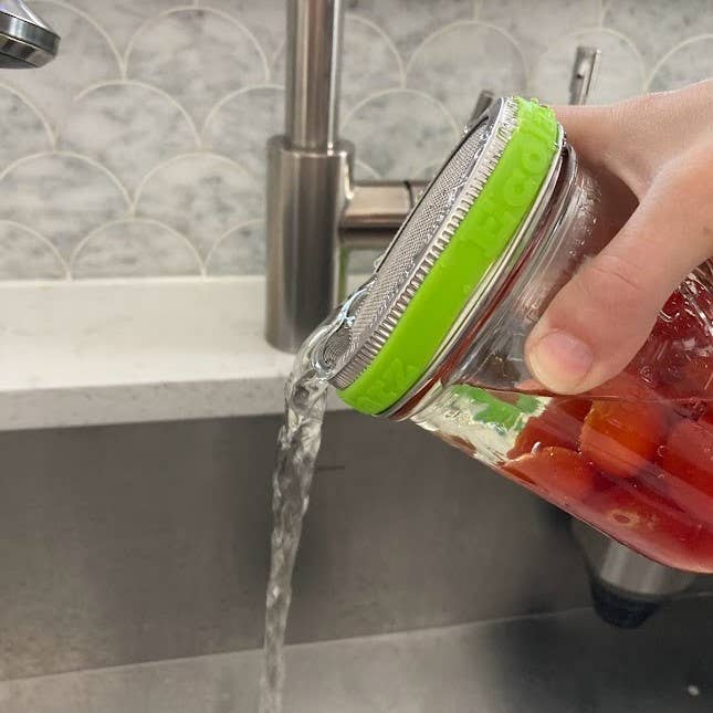 Person washing a jar with a green lid under running water in a kitchen sink.