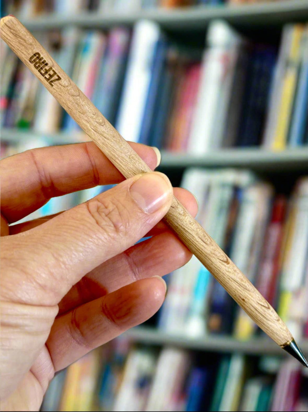 Hand holding a wooden everlasting pencil with a blurred bookshelf background