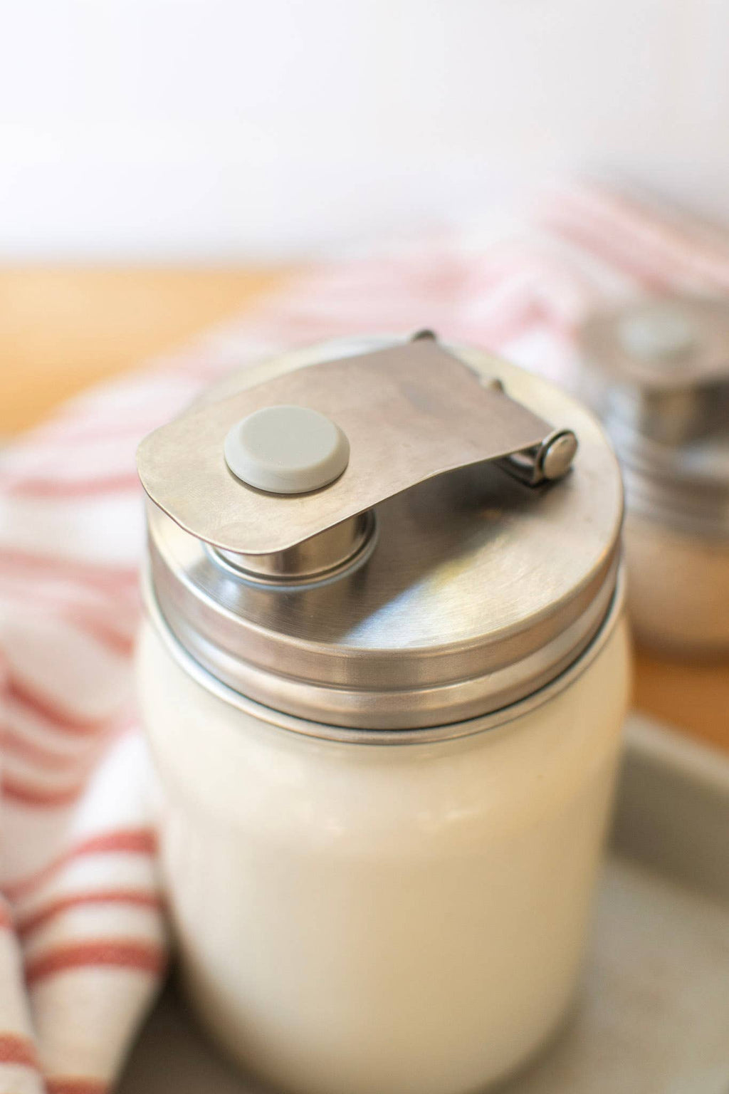 Stainless Steel pouring lid on a wide mouth jar with white liquid in it, on a white background on a wooden surface on a light plate.