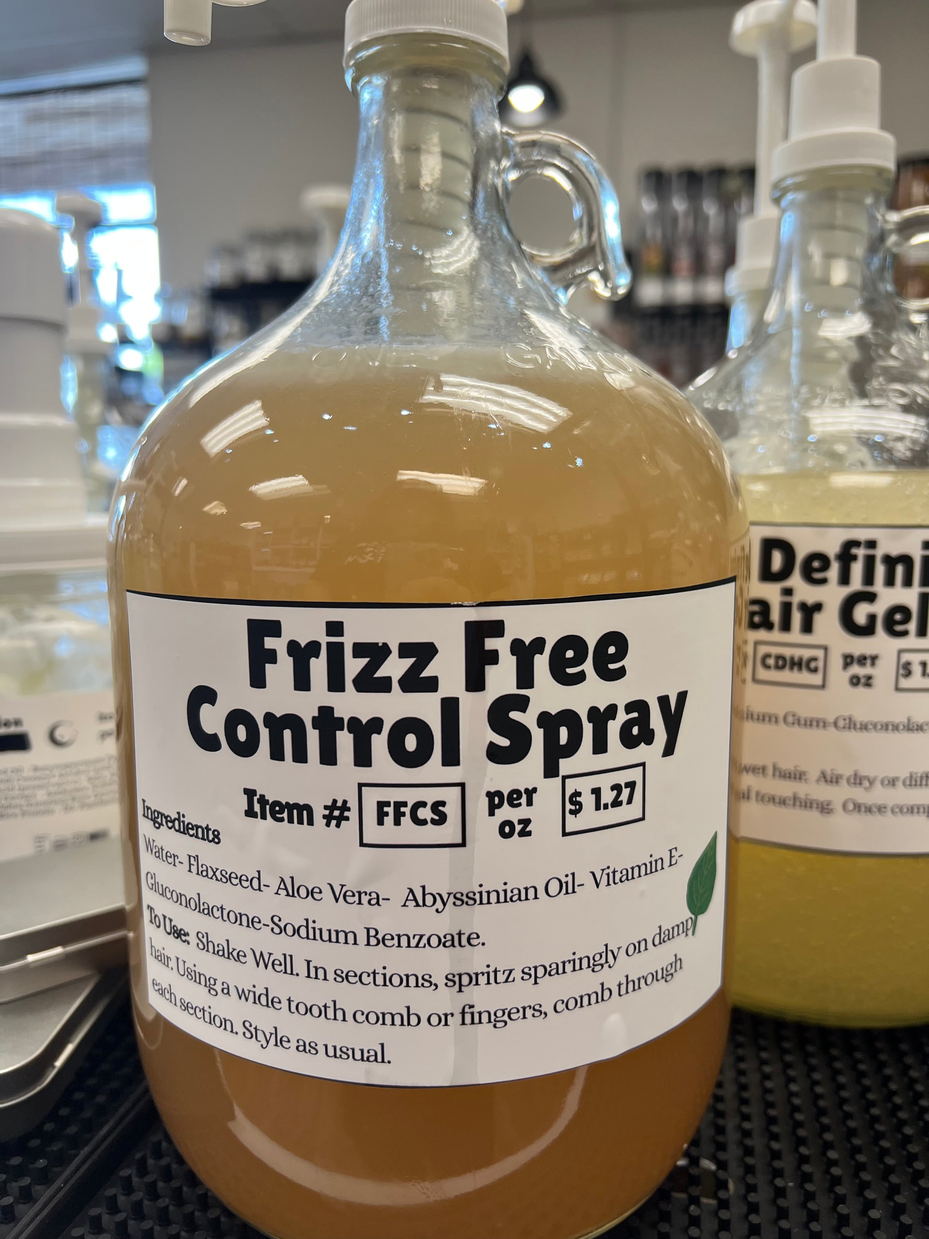 A one gallon glass pump jug of an amber colored liquid with frizz free label on a black table top, in front of other one gallon glass jugs.