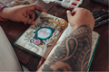 Person with tattooed arm holding a decorated notebook on a wooden surface