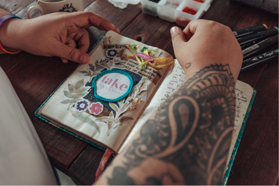 Person with tattooed arm holding a decorated notebook on a wooden surface