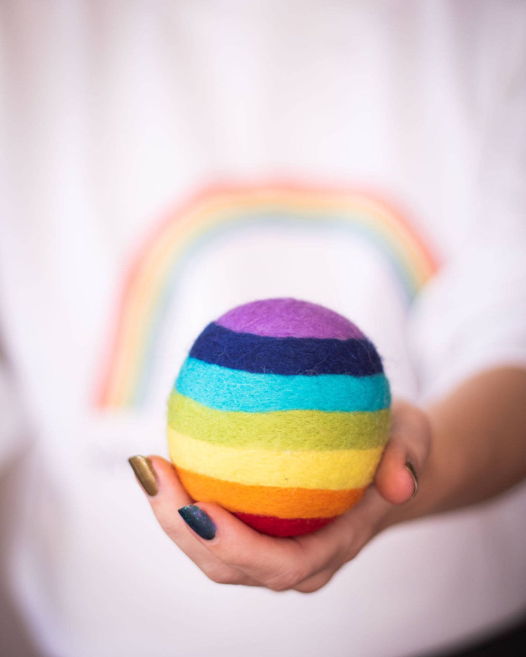 Hand holding a rainbow-colored wool dryer ball with a blurred rainbow arch in the background