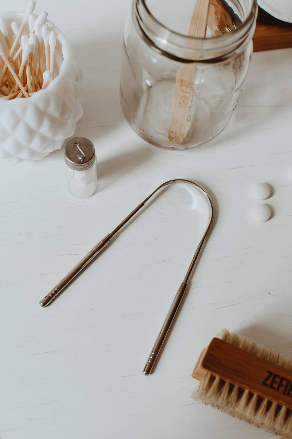 Clear glass jar with wooden stirrer, silver  tongue scraper and brush on a white surface.