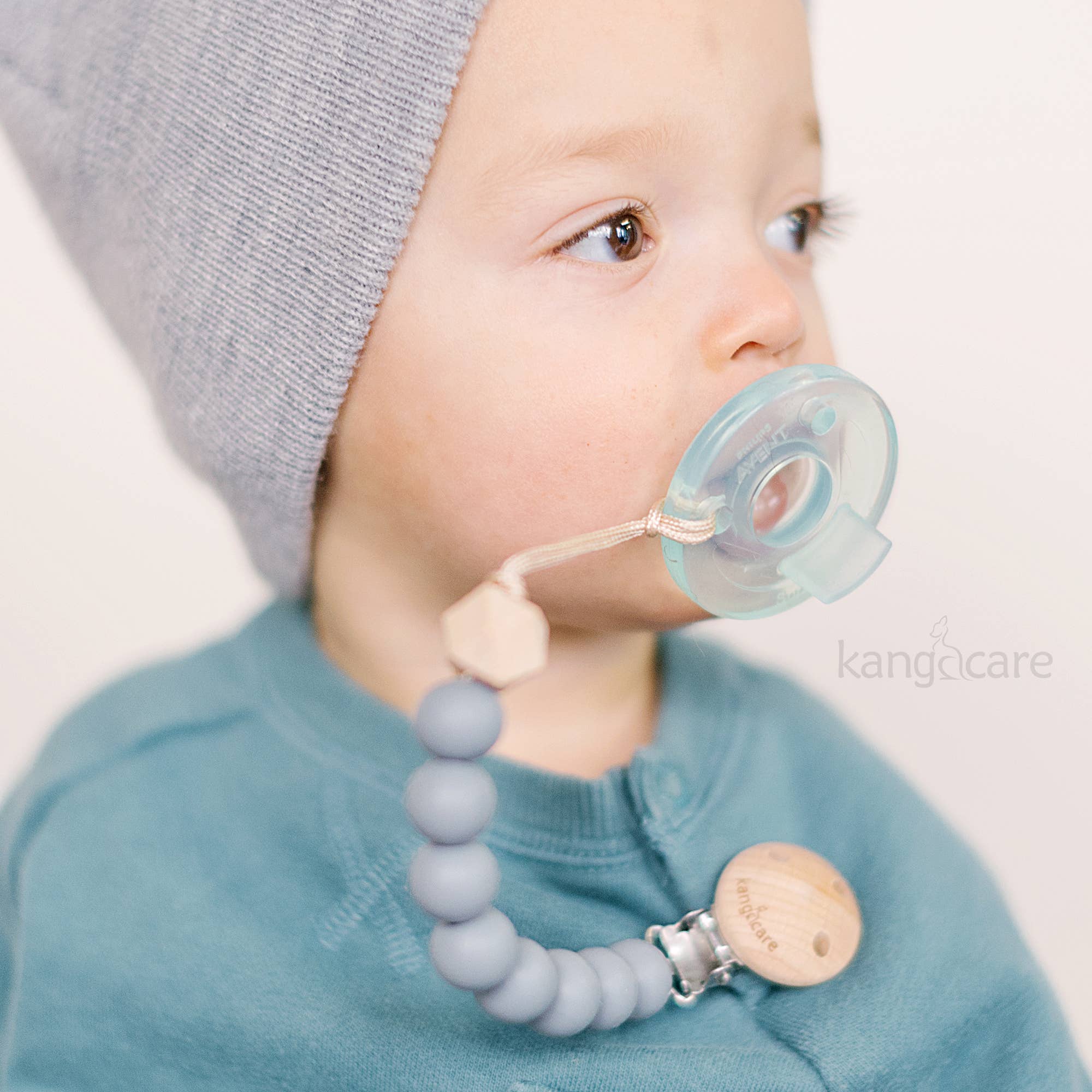 Baby wearing a gray knit hat and blue bib with a teething ring and pacifier holder.