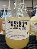 A one gallon glass pump jug of a light amber colored liquid with a curl defining gel label on a black table top, in front of other one gallon glass jugs.