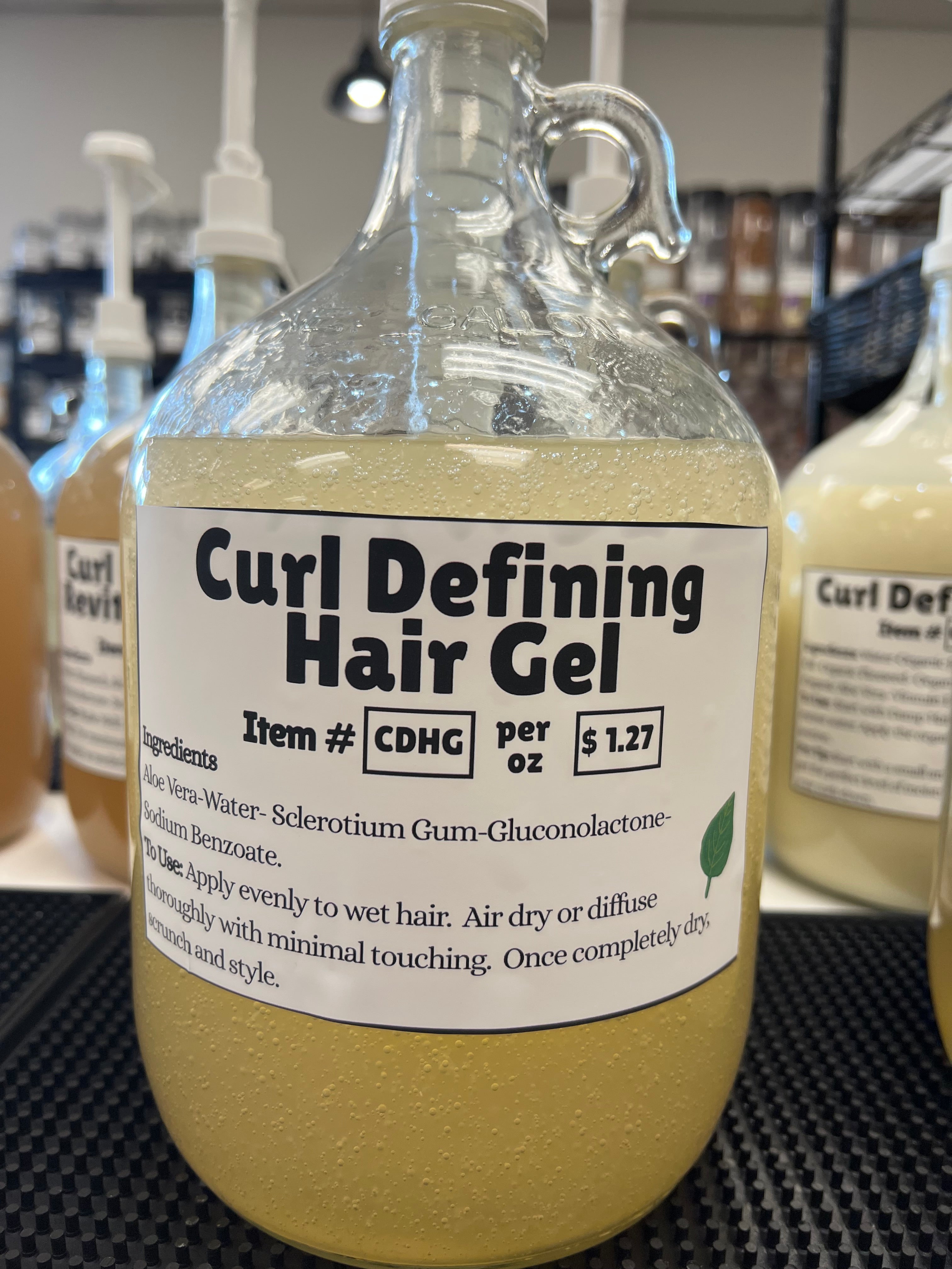 A one gallon glass pump jug of a light amber colored liquid with a curl defining gel label on a black table top, in front of other one gallon glass jugs.