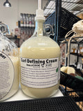 A one gallon glass pump jug of a light cream colored liquid with a curl cream label on a black table top, in front of other one gallon glass jugs.