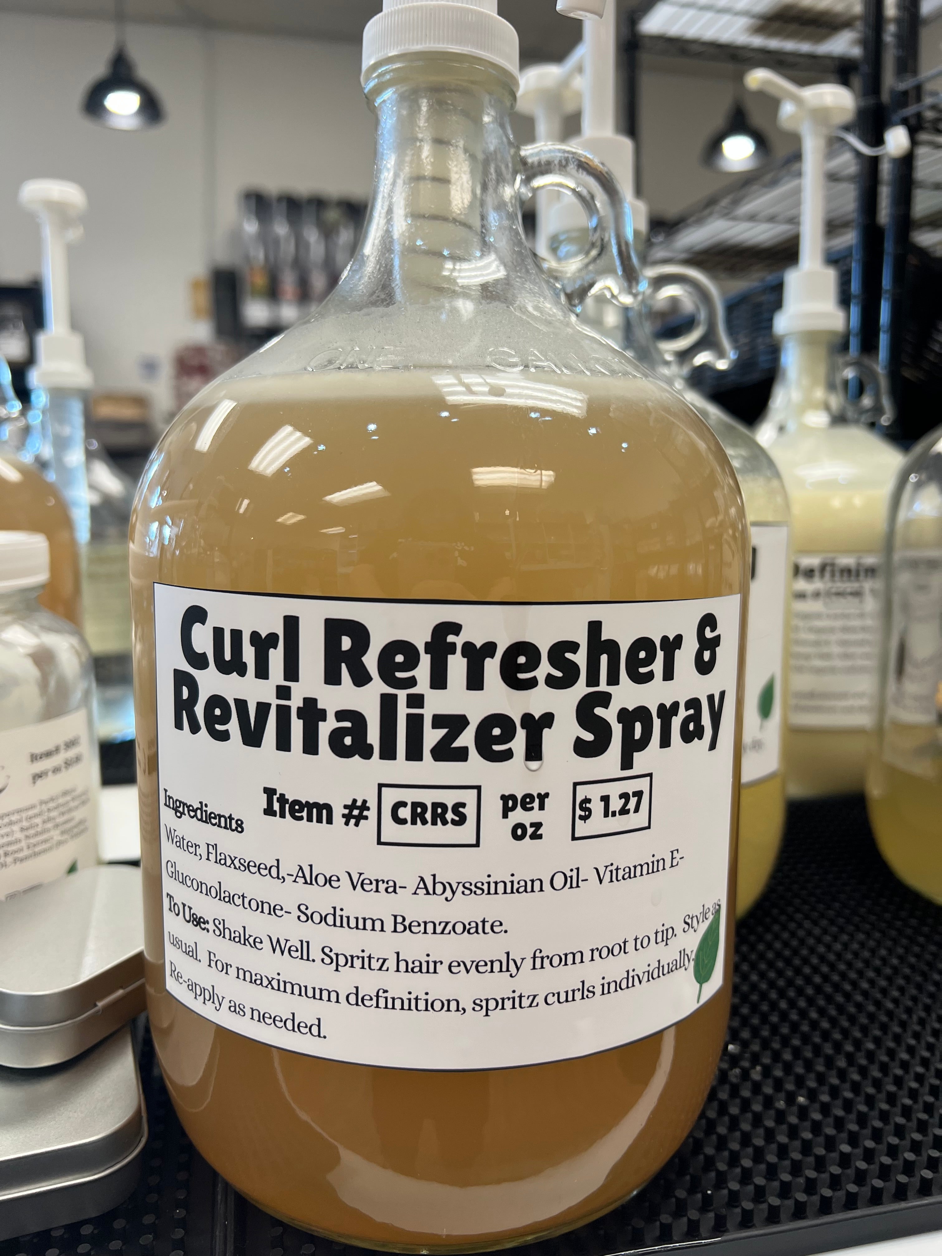 A one gallon glass pump jug of an amber colored liquid with a curl refresh relabel on a black table top, in front of other one gallon glass jugs.