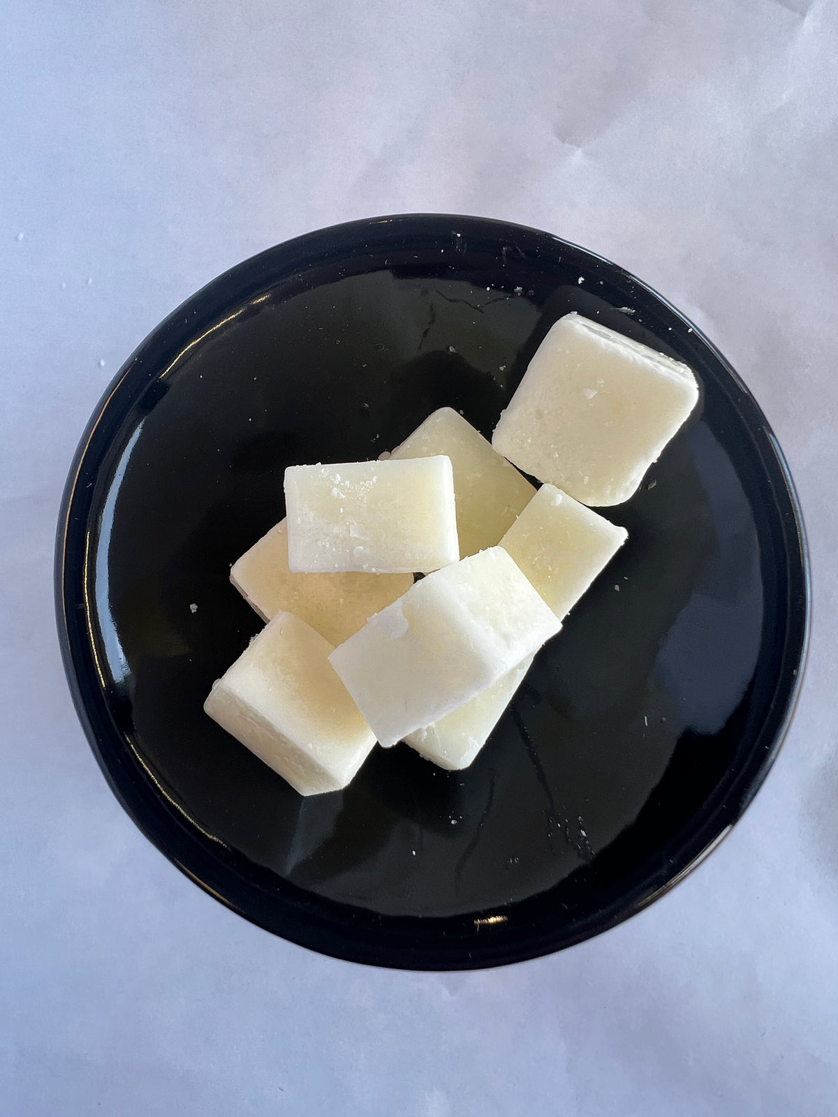 Several pale cream-colored wax melts arranged on a glossy black plate, photographed from above against a light gray background.
