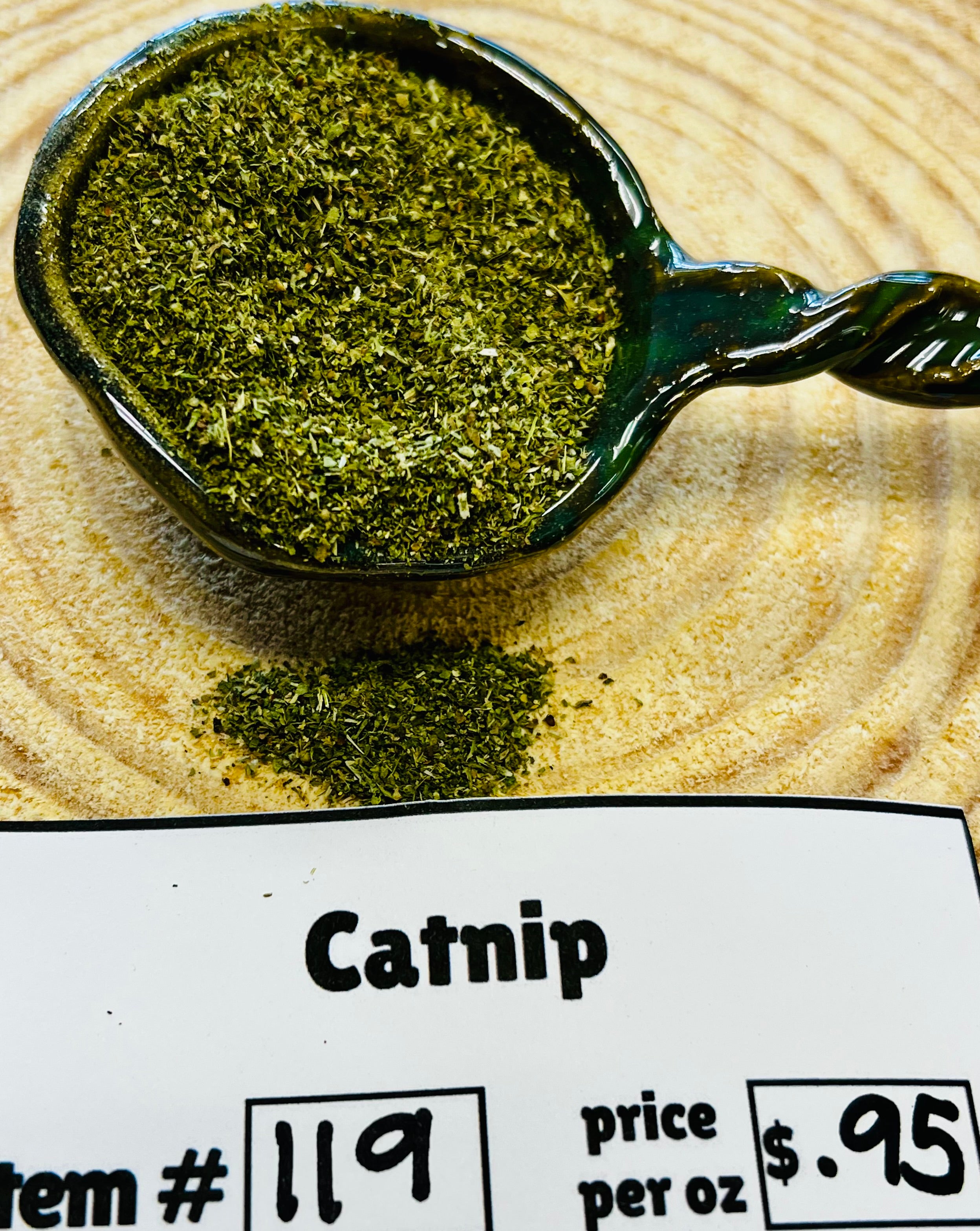Spoonful of catnip on a wooden surface with a label indicating the item number, price per ounce, and product name.