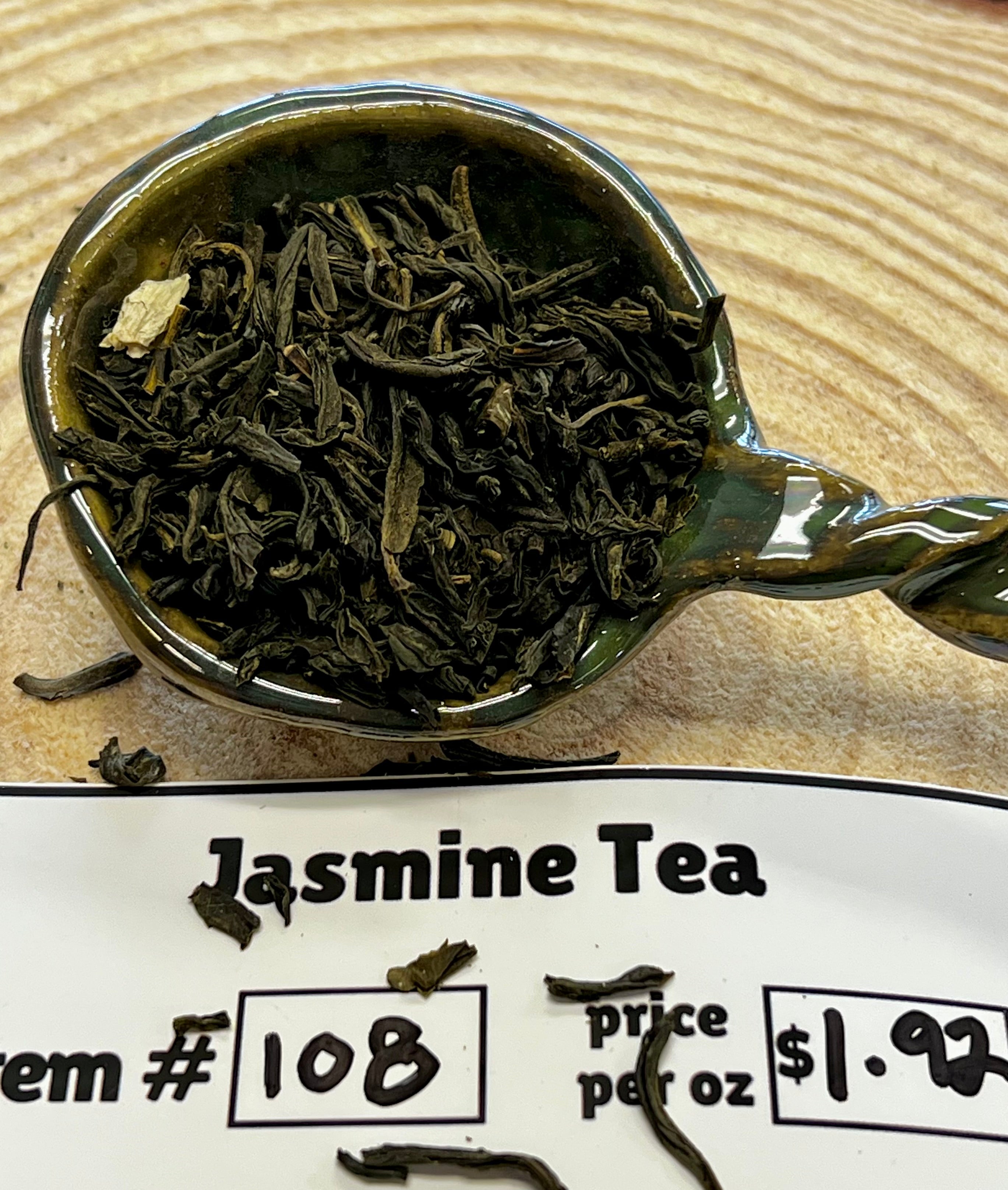Tea leaves in a metal scoop with a label indicating 'Jasmine Tea' and pricing.
