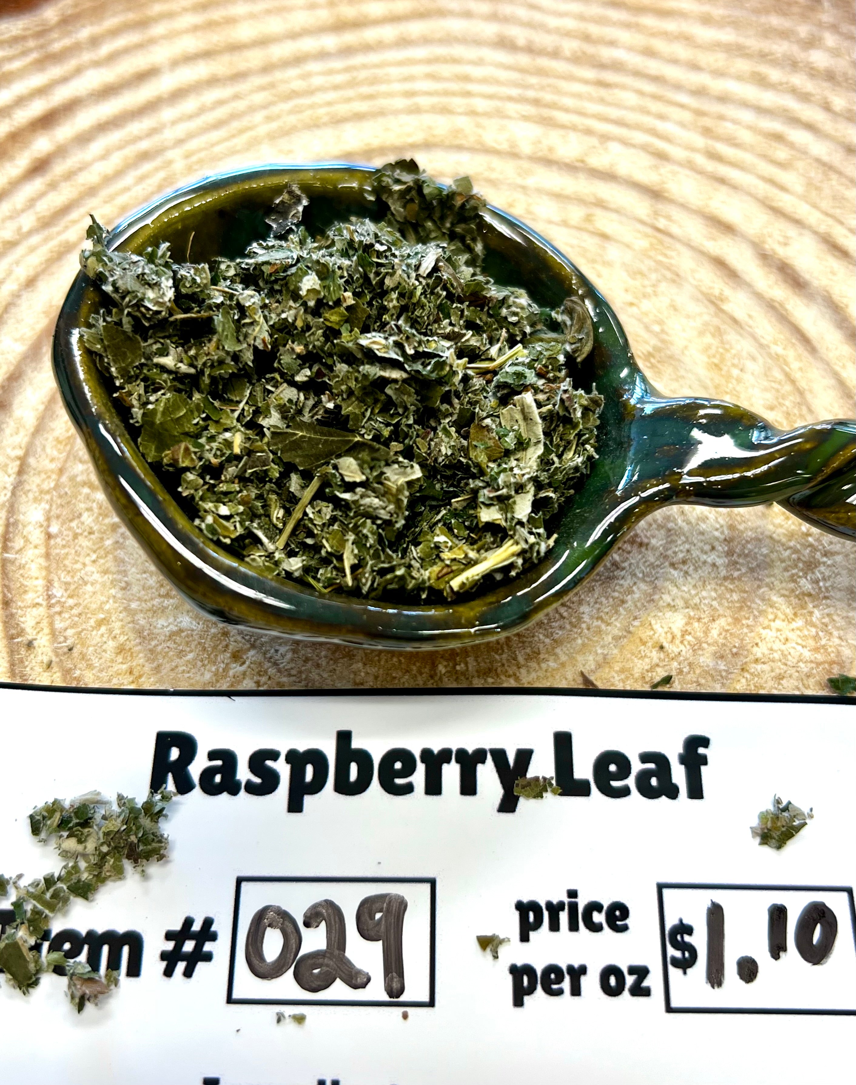 Teaspoon filled with dried raspberry leaf on a wooden surface, with text indicating item number and price.