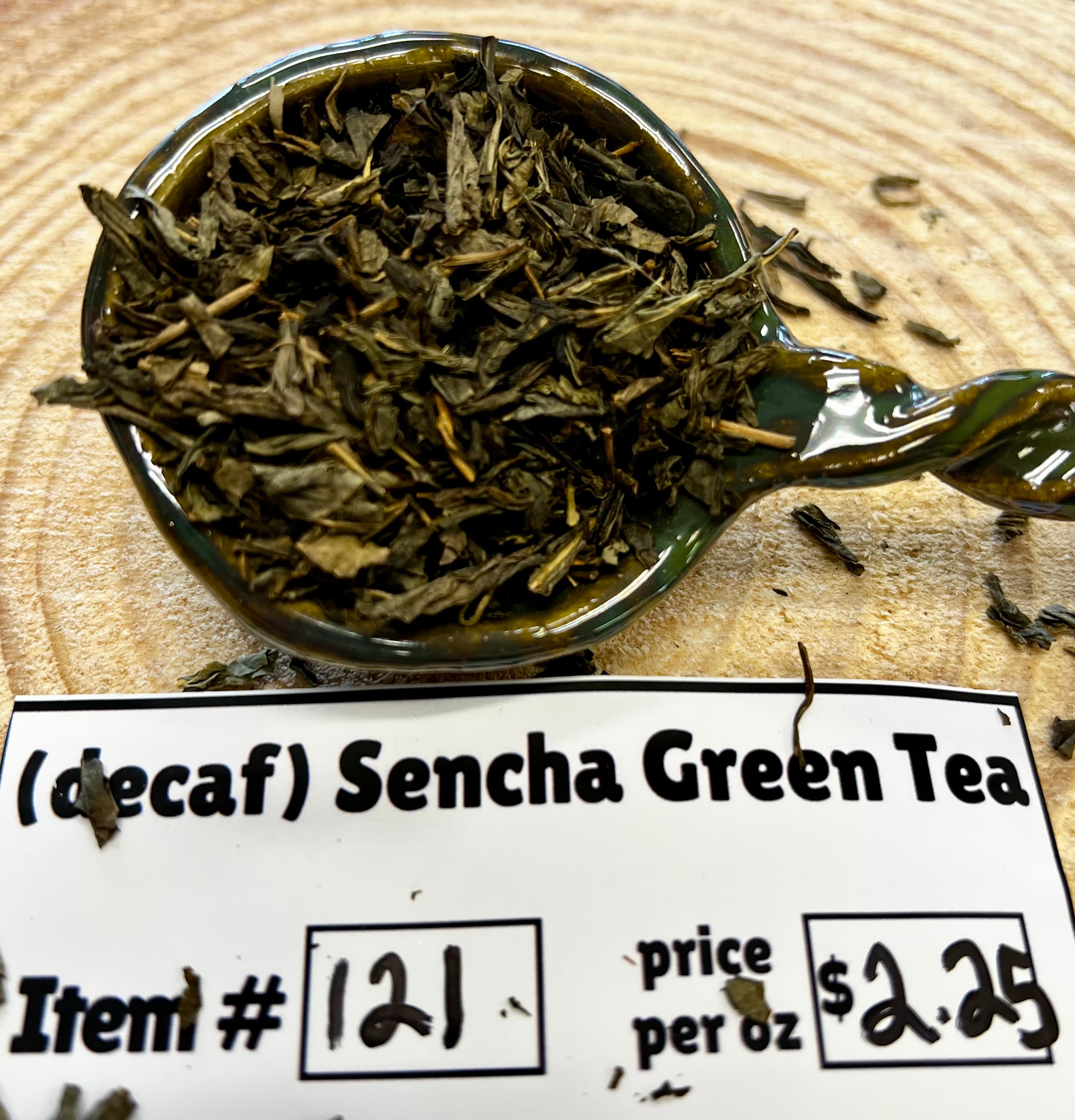 Spoon filled with green tea leaves on a wooden surface with a label below.