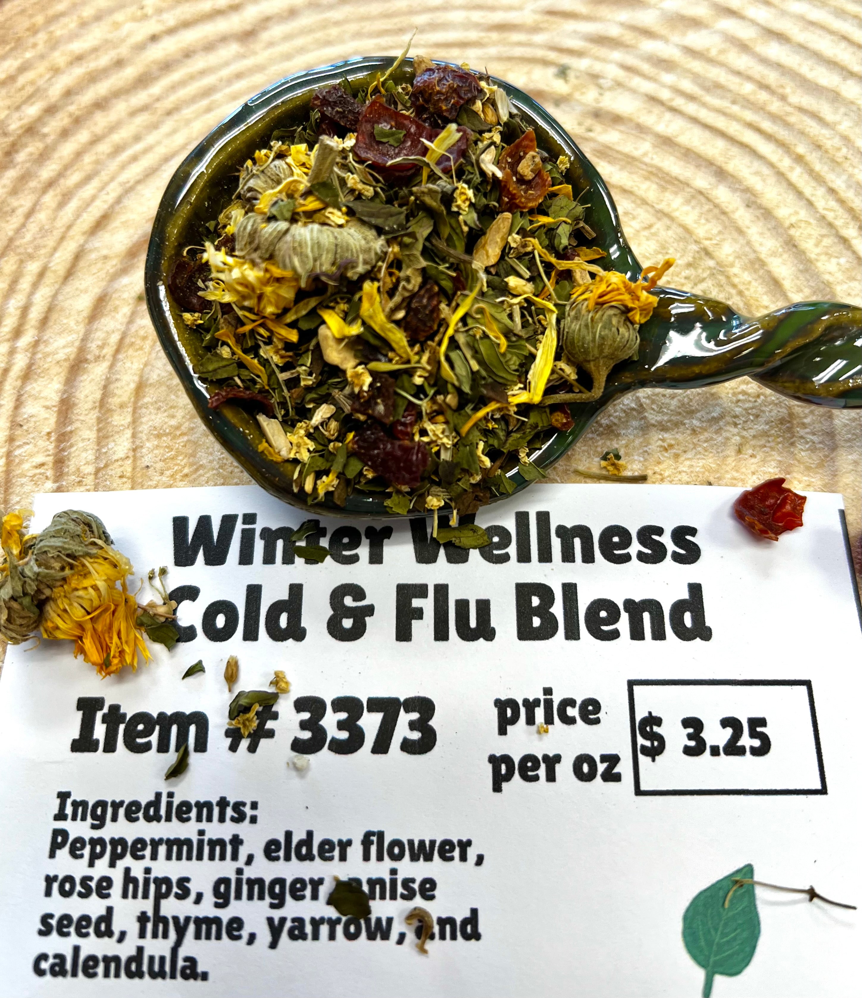Tea blend with botanicals in a spoon with a label for 'Winter Wellness Cold & Flu Blend' on a wooden surface.
