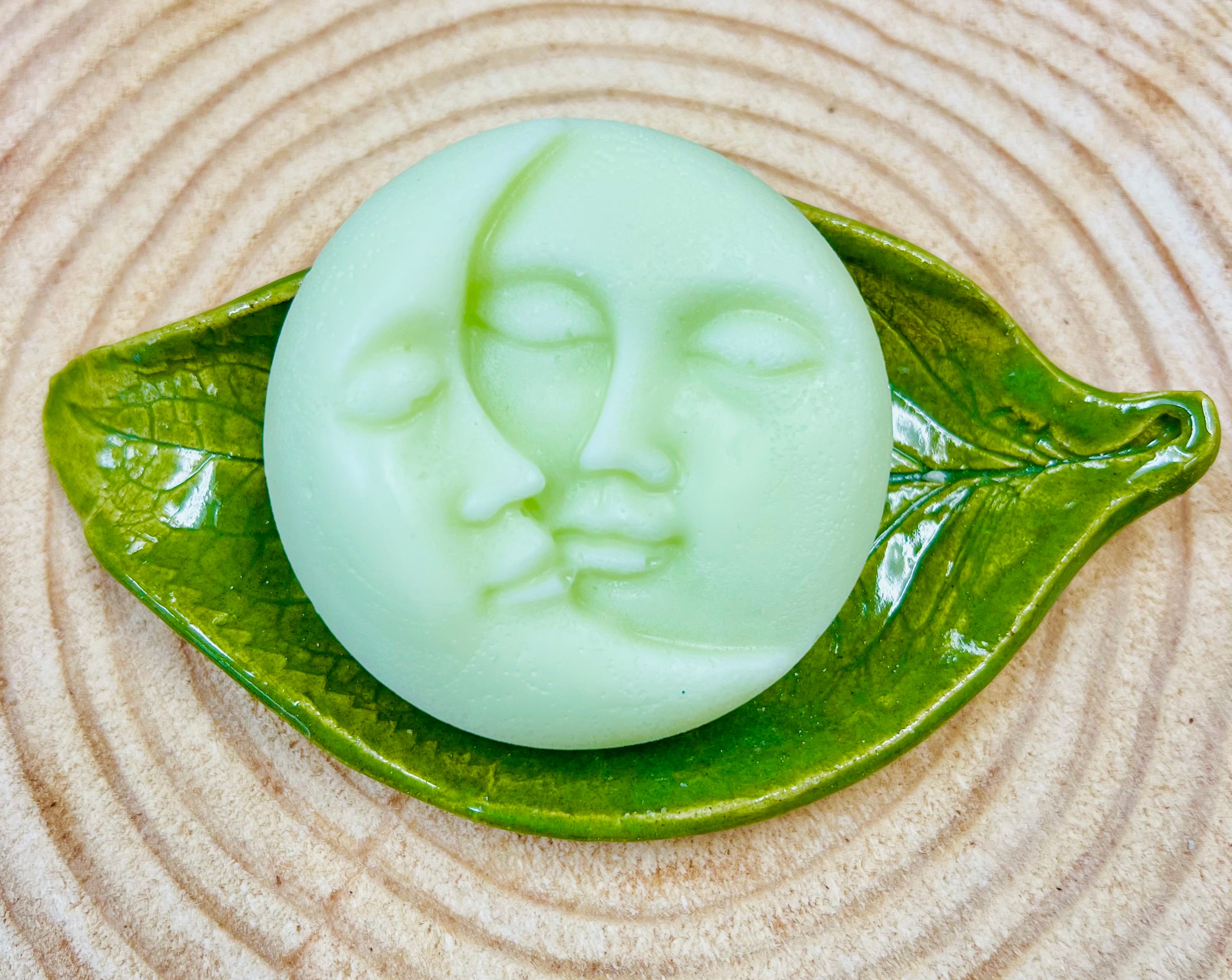 Green round conditioner bar with moon & Sun face designs on a ceramic on a leaf on a wood surface.