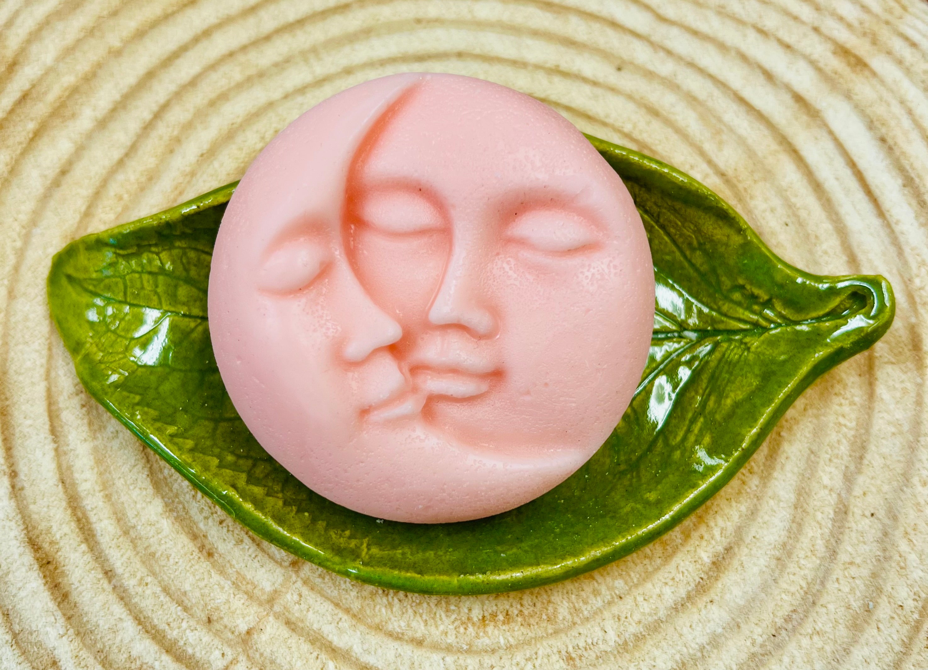 Pink round conditioner bar with moon & Sun face designs on a ceramic on a leaf on a wood surface.