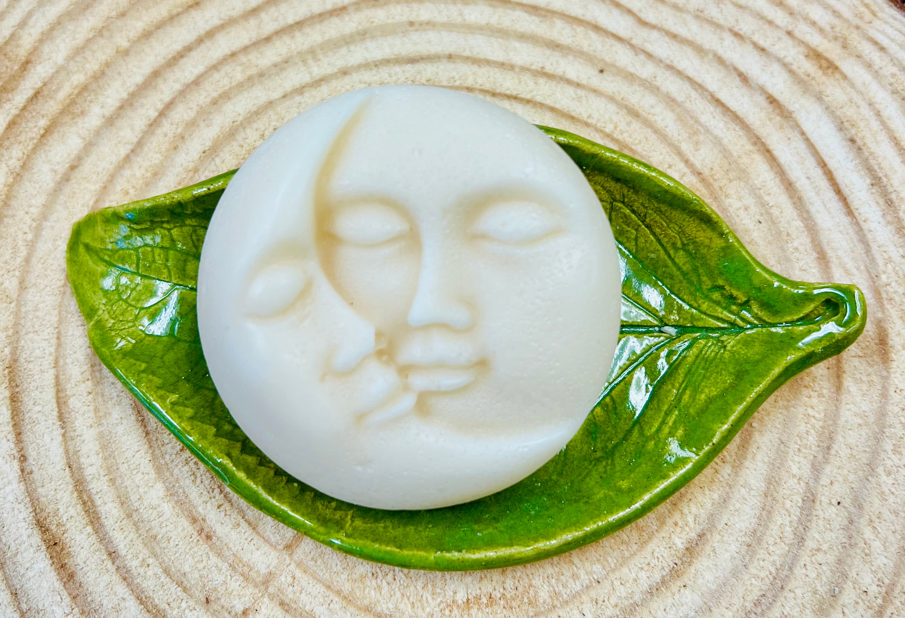 White round conditioner bar with moon & Sun face designs on a ceramic on a leaf on a wood surface.