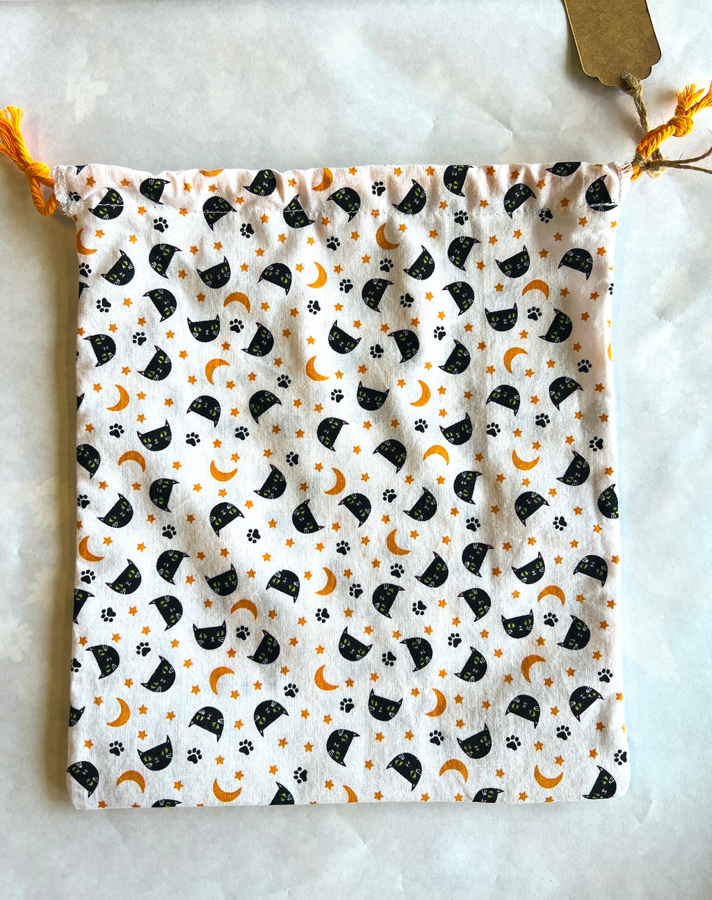 Fabric drawstring bag laying flat with black cat face, orange crescent moons, and black paw prints on a white background with bright orange cording. 