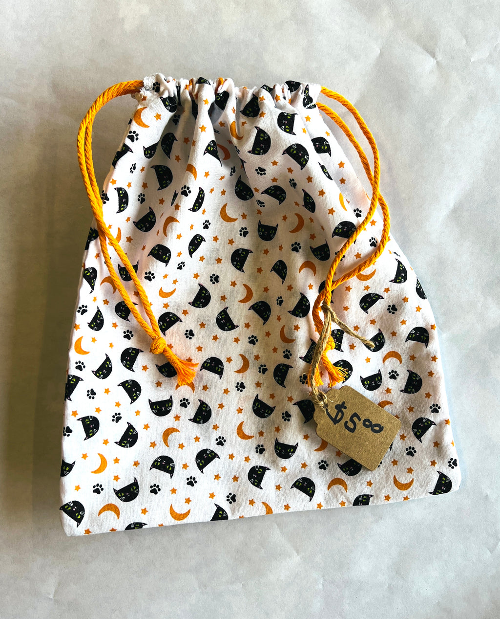 A small drawstring pouch with a white fabric body printed with black cat silhouettes, orange crescent moons and paw prints, and bright orange drawstring cords. A small kraft paper tag hangs from one cord.