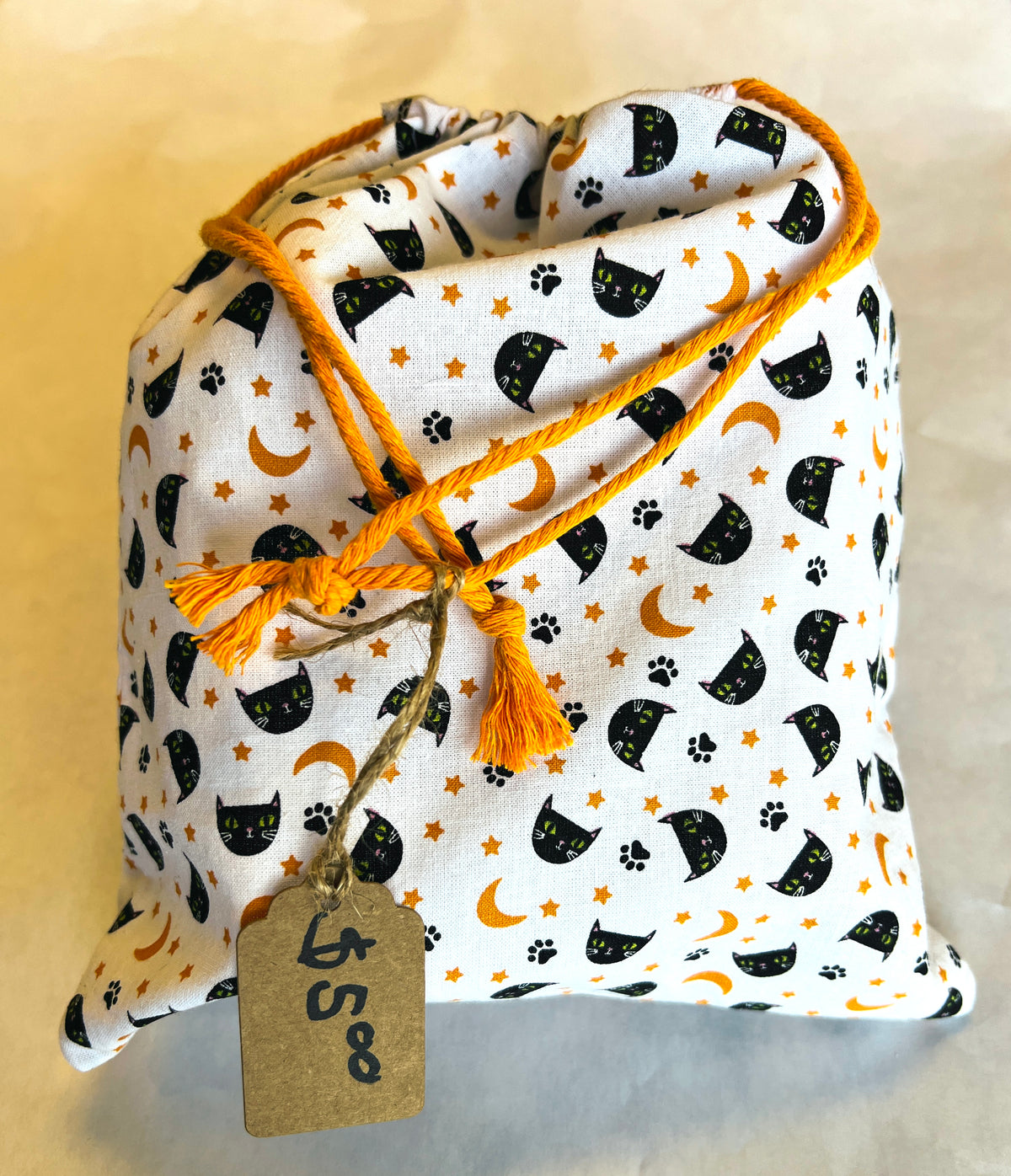 A fabric drawstring pouch with a white background printed with black cat silhouettes, orange crescent moons, and orange paw prints, featuring orange drawstrings and a hanging paper price tag.