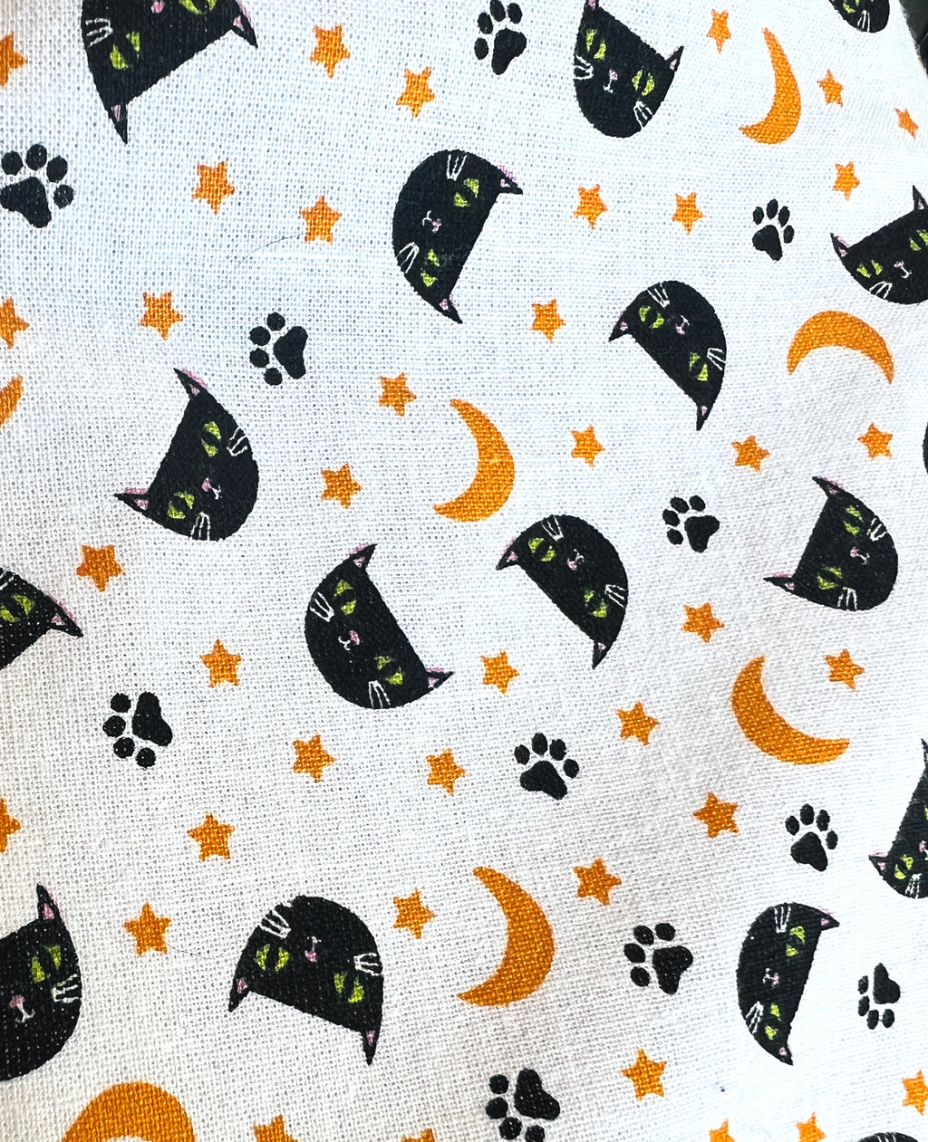 A close-up of a white fabric with a repeating pattern of black cat heads in crescent moon shapes, orange crescent moons, orange stars, and black paw prints.