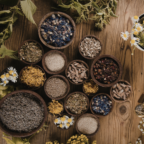 Herbs and flowers in small wooden bowls on a wooden surface with green leaves and white flowers.