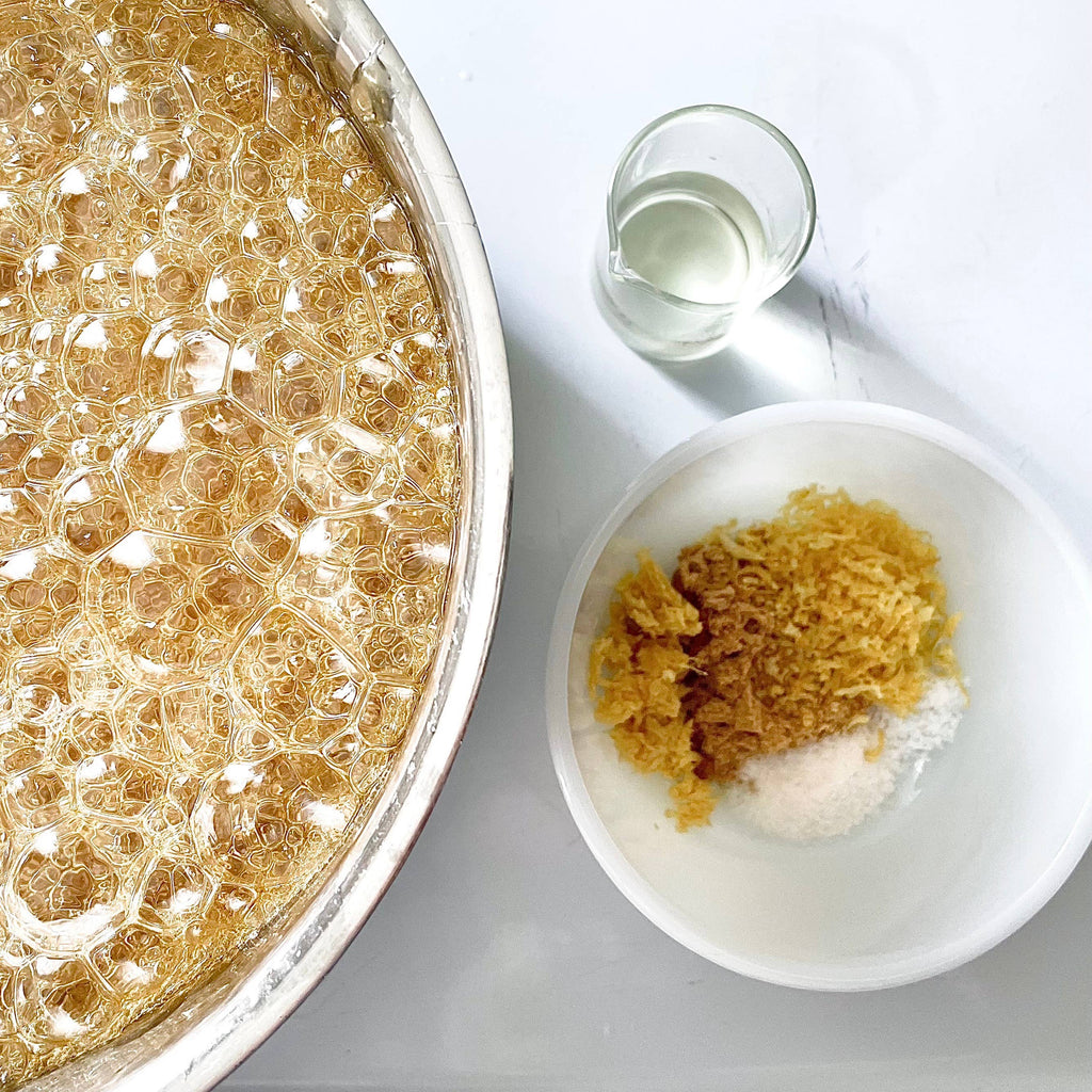 A close-up overhead view of a large metal bowl filled with golden amber honey or syrup, alongside a small glass of clear liquid and a white bowl containing crushed golden material and white powder on a light surface.