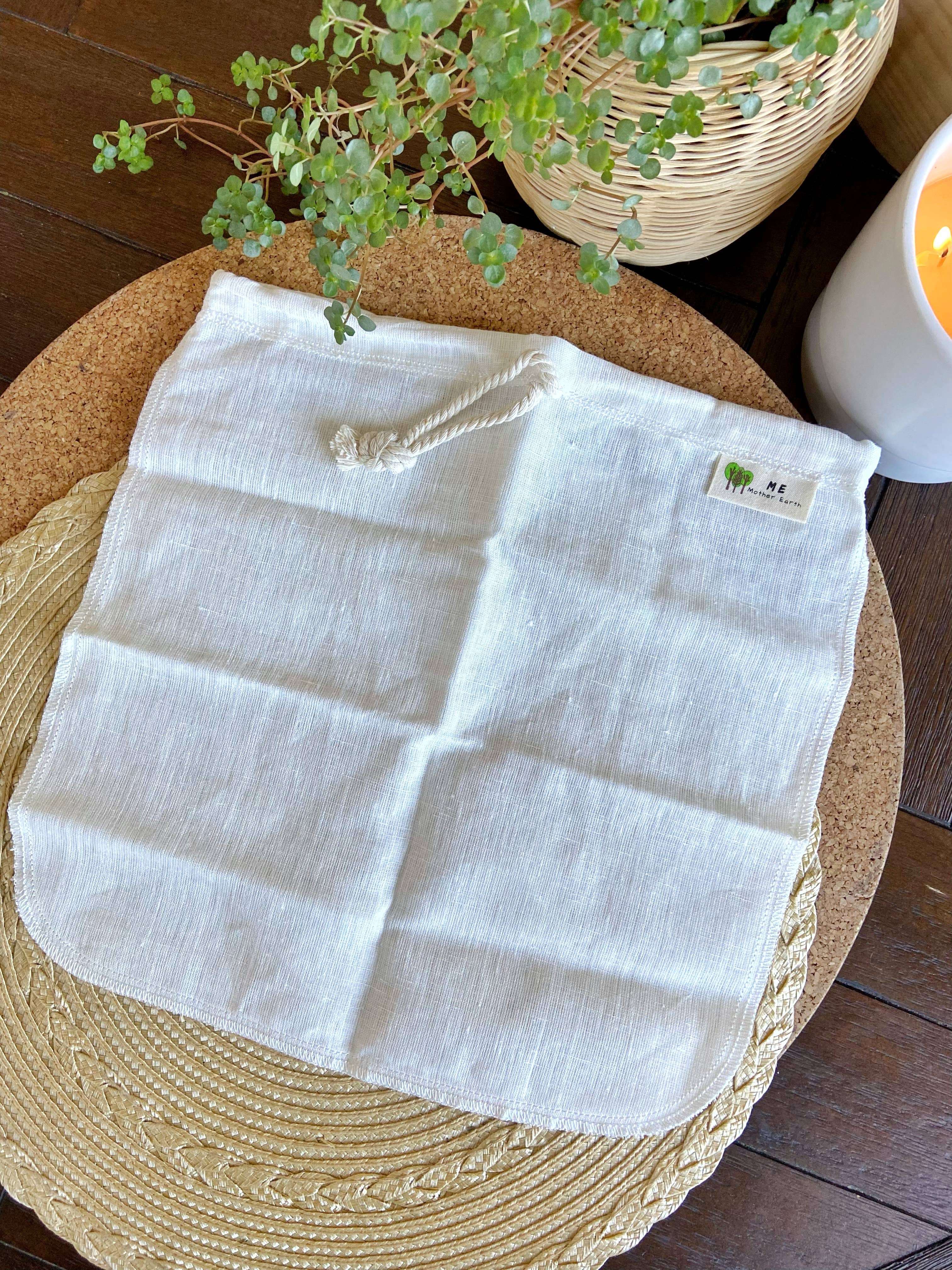 An off-white linen cloth pouch with a rope handle, laid flat on a round woven mat. The background features a wooden floor, a potted green plant in a wicker basket, and a white candle.