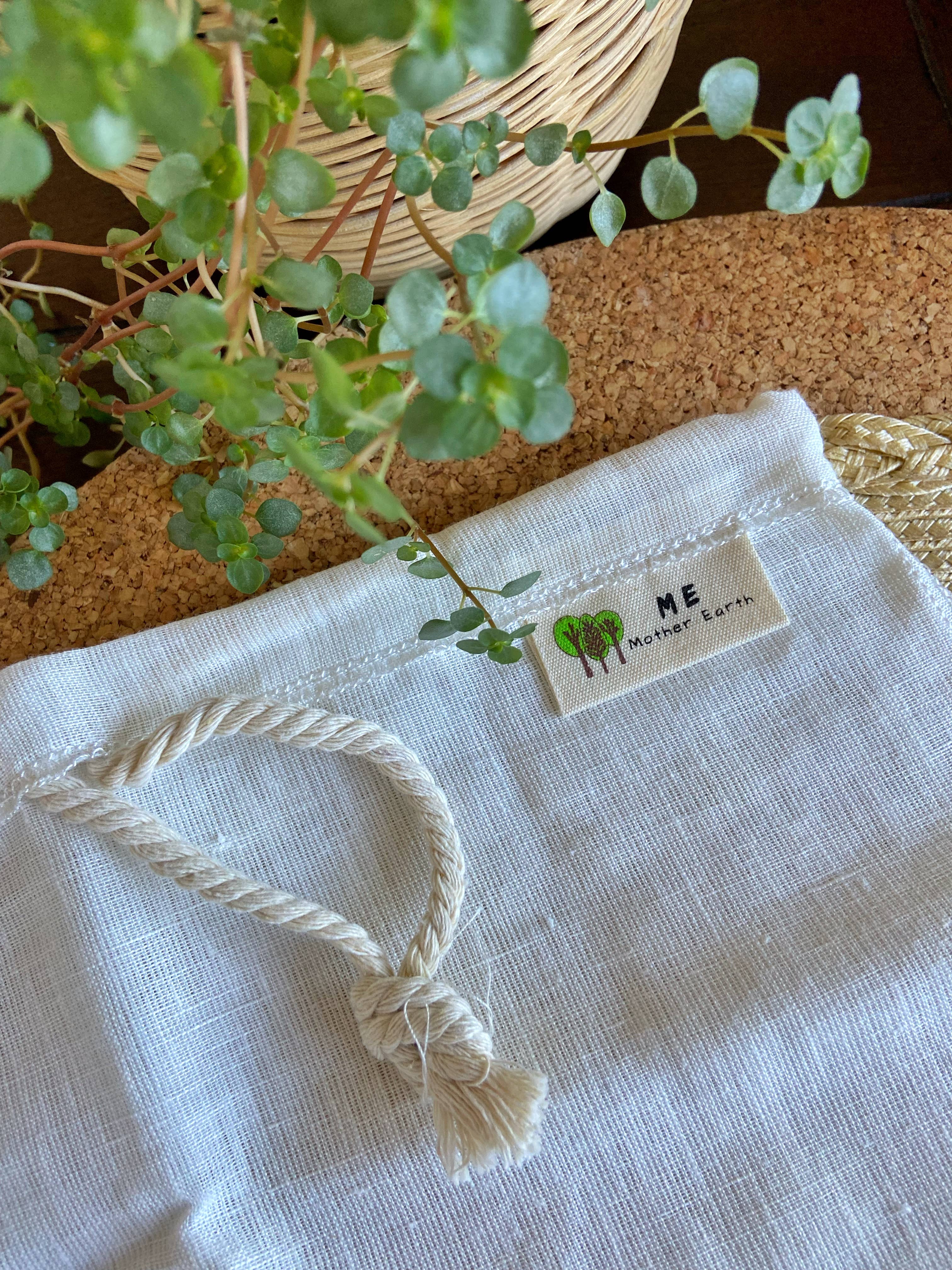 A close-up of a white textured fabric pouch or bag with a braided rope tassel and a sewn-on label reading 'ME Mother Earth' with a small tree graphic, styled with green leaves and a cork surface.