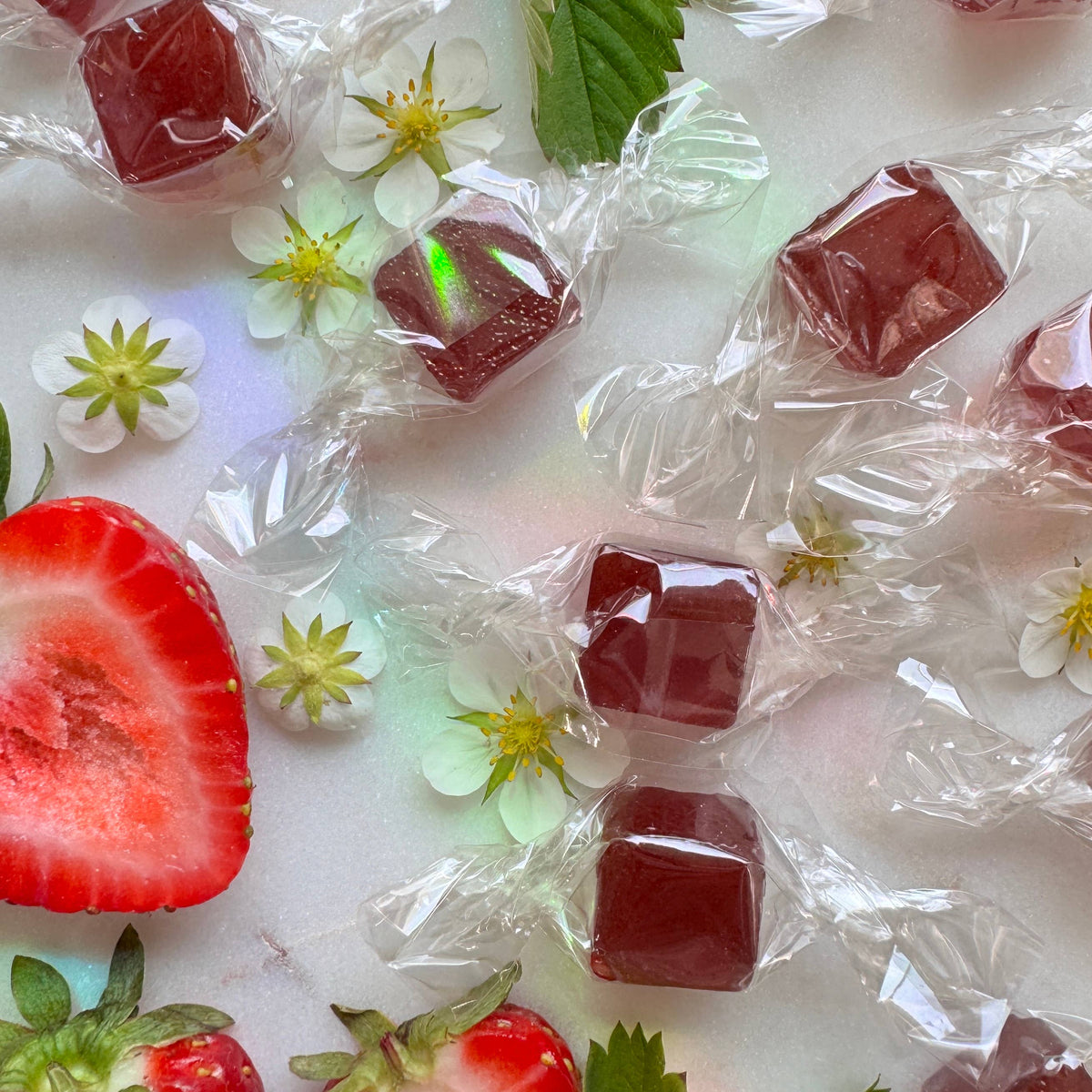 Red jelly cubes in clear wrappers on a white surface with strawberries and flowers.