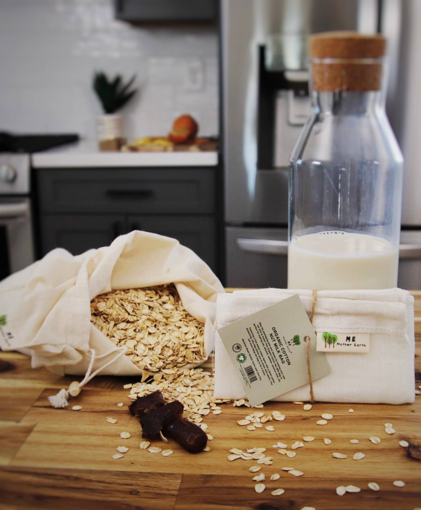 A styled kitchen scene showing a cloth bag filled with rolled oats next to a glass bottle of milk and a folded cloth towel with a tag, on a wooden countertop.