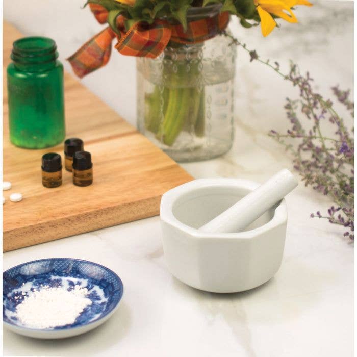 Mortar and pestle on a table with bottles, a cutting board, and flowers in the background.
