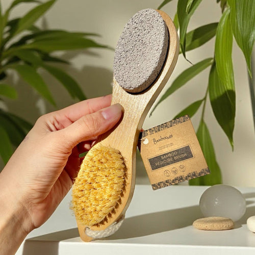 Hand holding a bamboo foot scrub brush with a label, against a natural background with plants.
