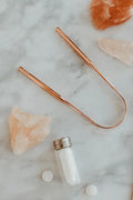 Rose gold metal  tongue scraper on a marble surface with salt container and crystals.