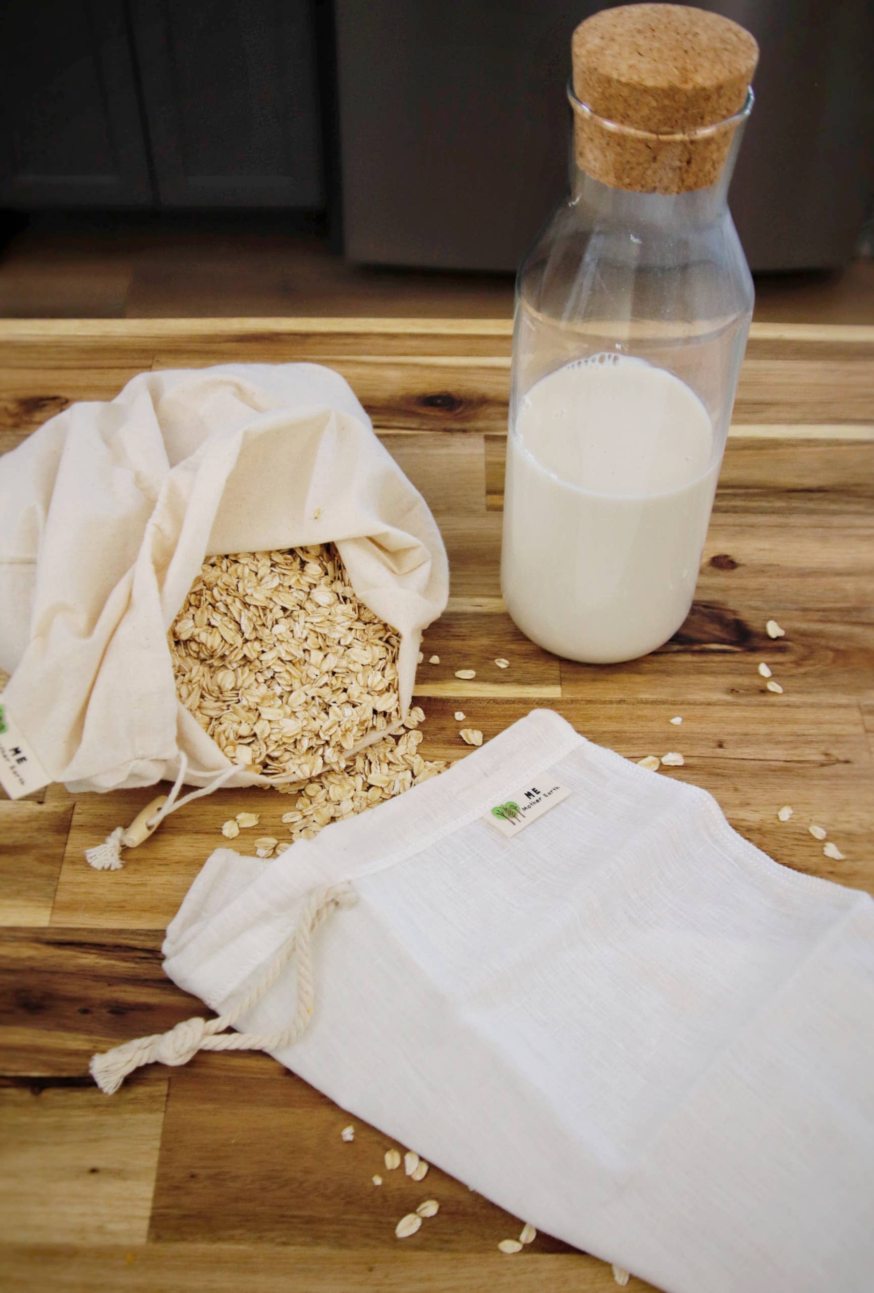 Two reusable cotton drawstring bags, one filled with oats and one empty, displayed on a wooden surface next to a glass bottle with a cork stopper containing milk.