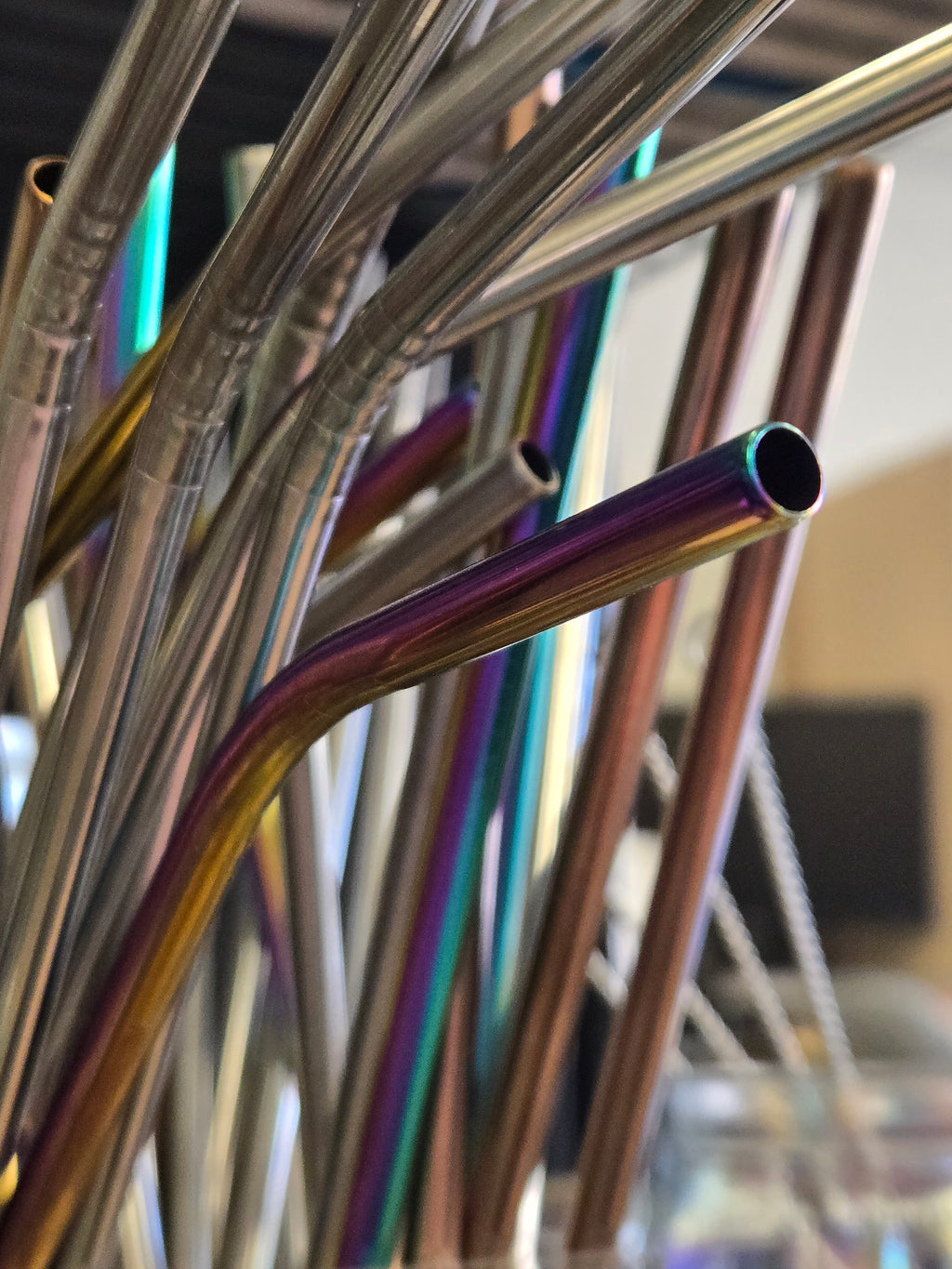 A close-up of a curved stainless steel straw with an iridescent rainbow finish, surrounded by other metallic straws in a cluster.