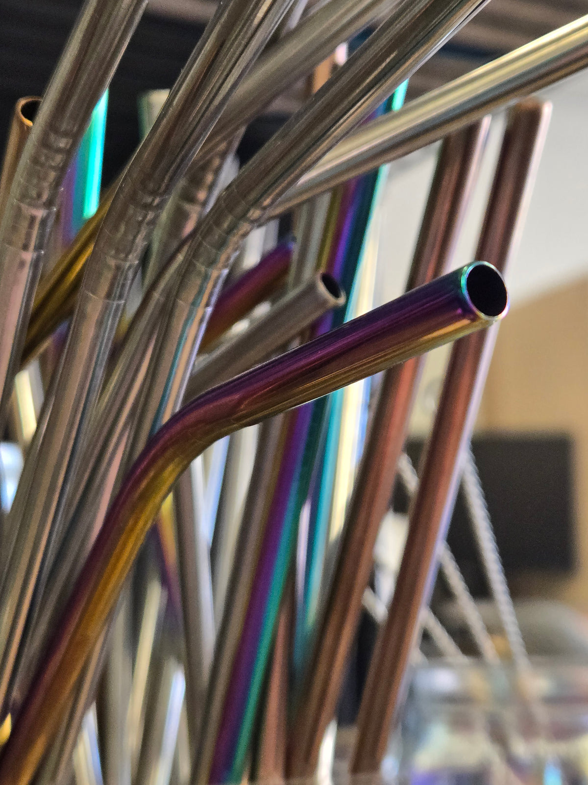 A close-up of a curved stainless steel straw with an iridescent rainbow finish, surrounded by other metallic straws in a cluster.