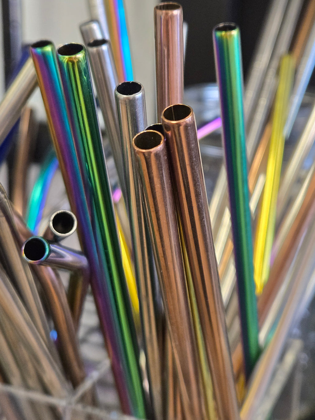 A close-up of multiple reusable metal straws with an iridescent rainbow finish and some metallic copper-toned straws, standing in a holder.