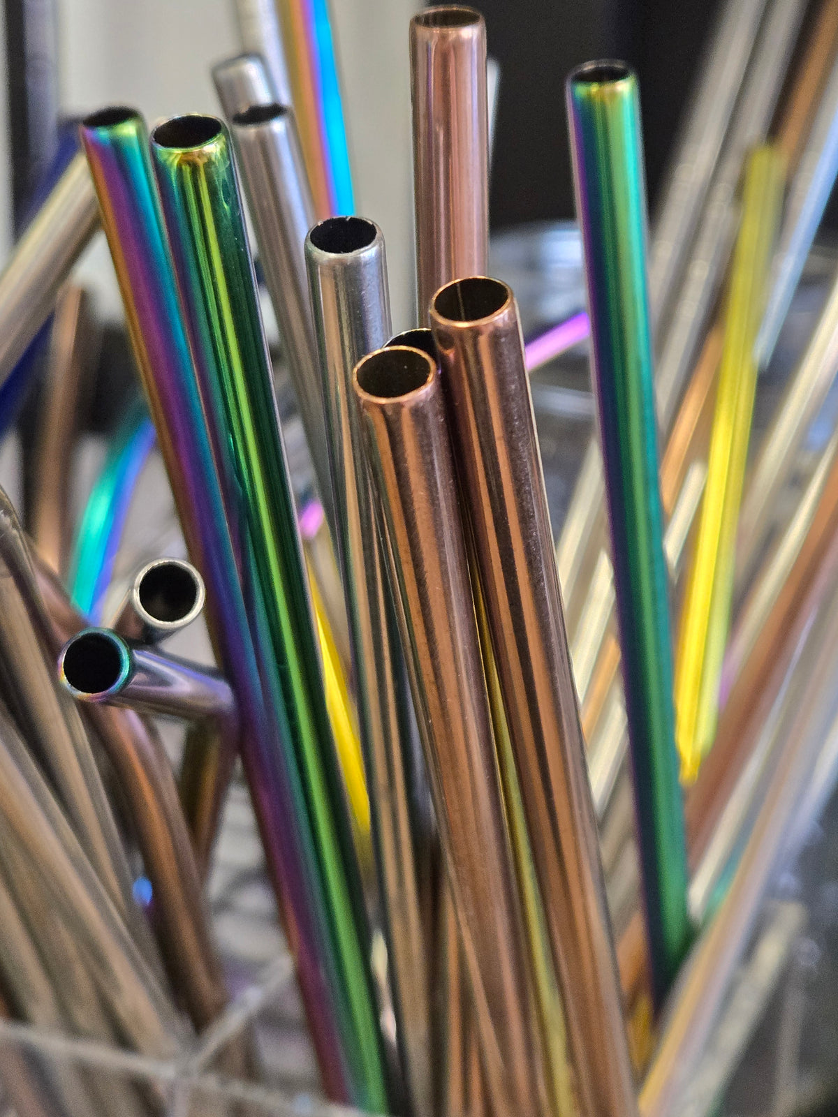 A close-up of multiple reusable metal straws with an iridescent rainbow finish and some metallic copper-toned straws, standing in a holder.